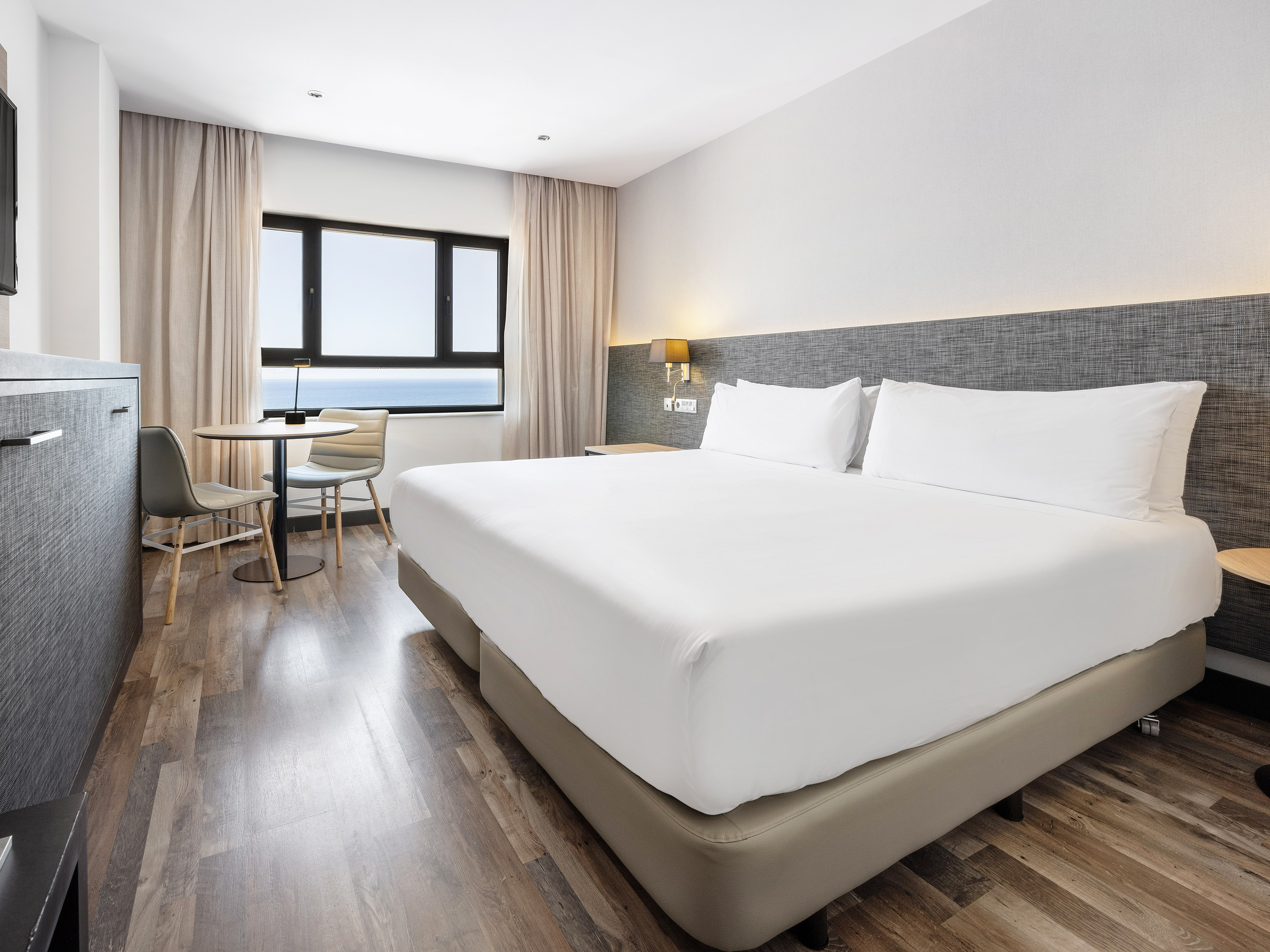 Modern hotel bedroom with king bed, ocean view, and wooden floor.