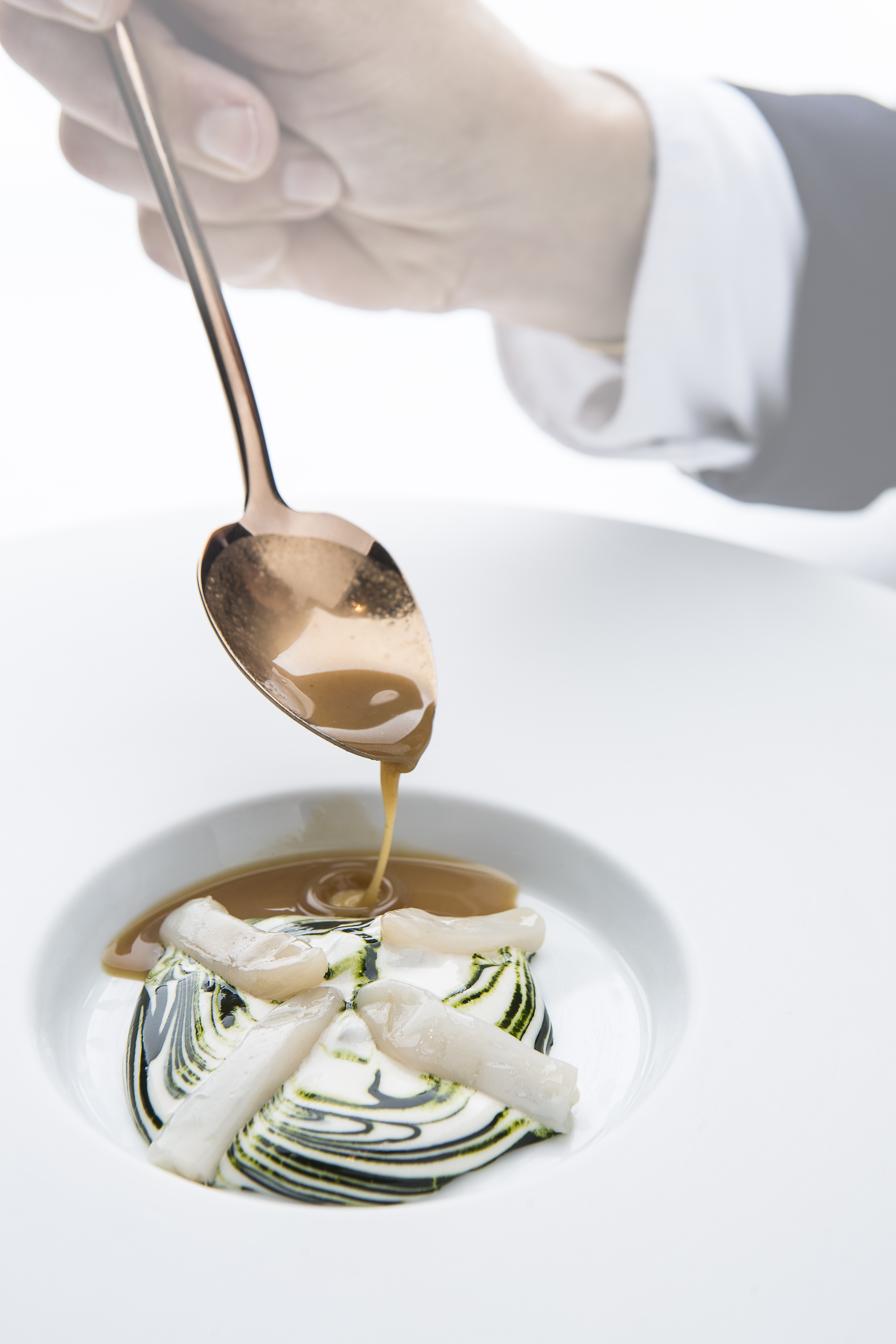 Chef's hand pouring brown sauce over gourmet seafood on artistic swirled plate.