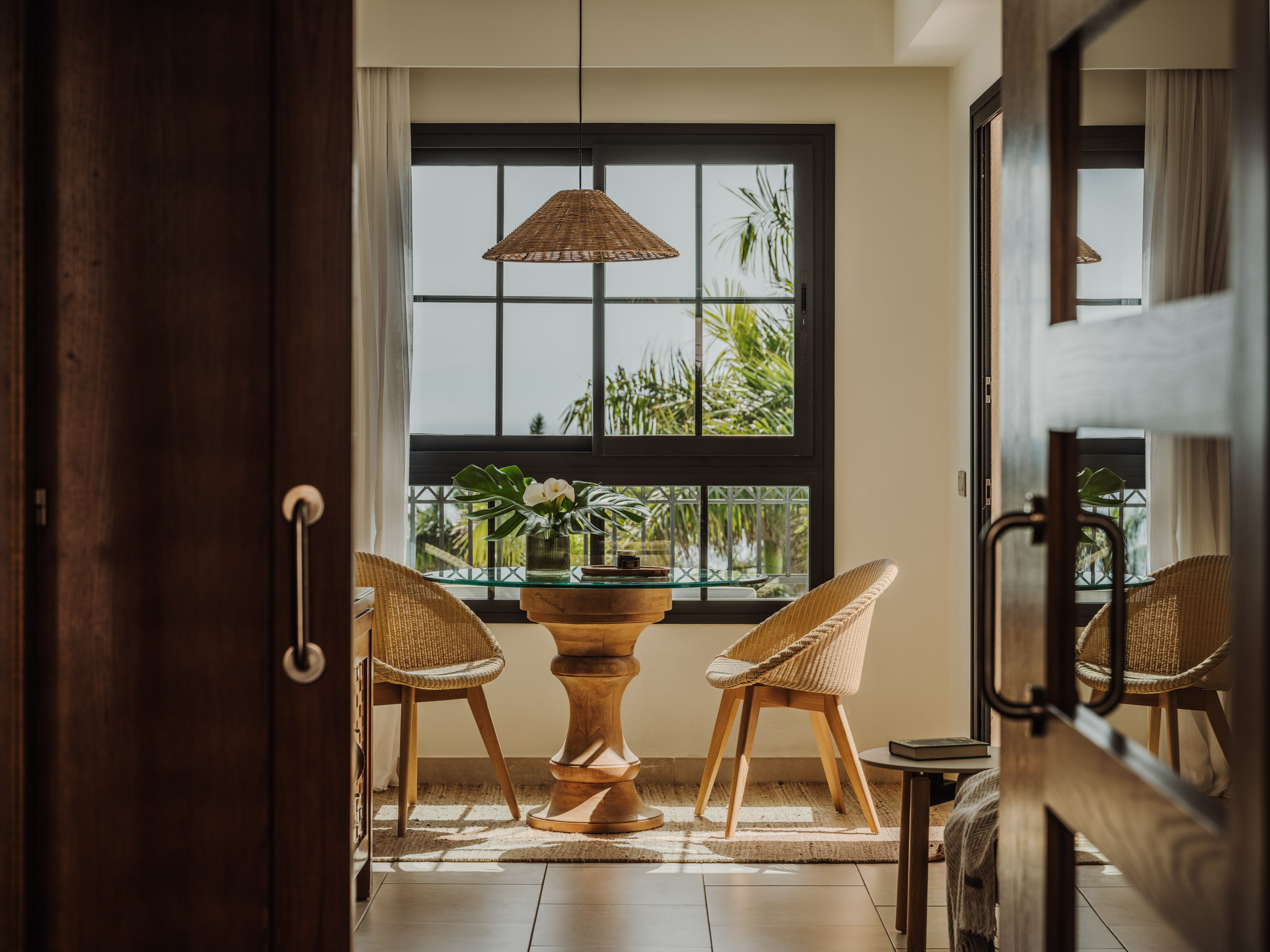 Tropical dining area with wicker chairs, wooden table, window and palm trees.