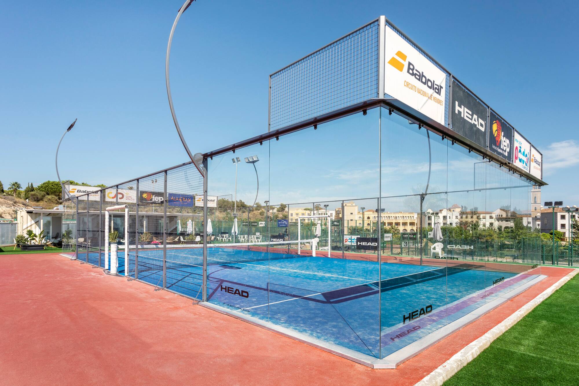 Blue outdoor padel court with glass walls, red surrounds, and Babolat/HEAD branding.