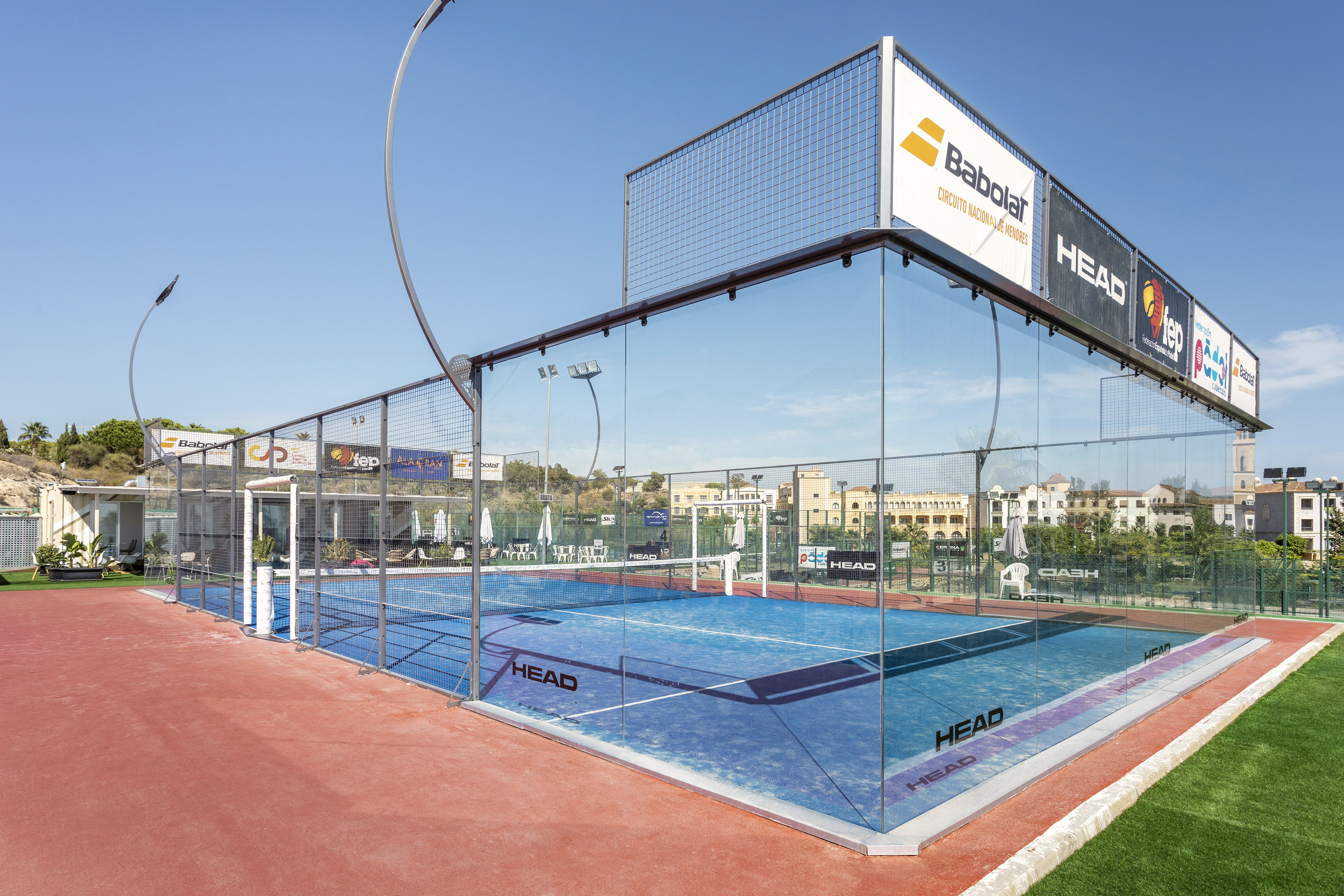 Blue outdoor padel court with glass walls, red surrounds, and Babolat/HEAD branding.