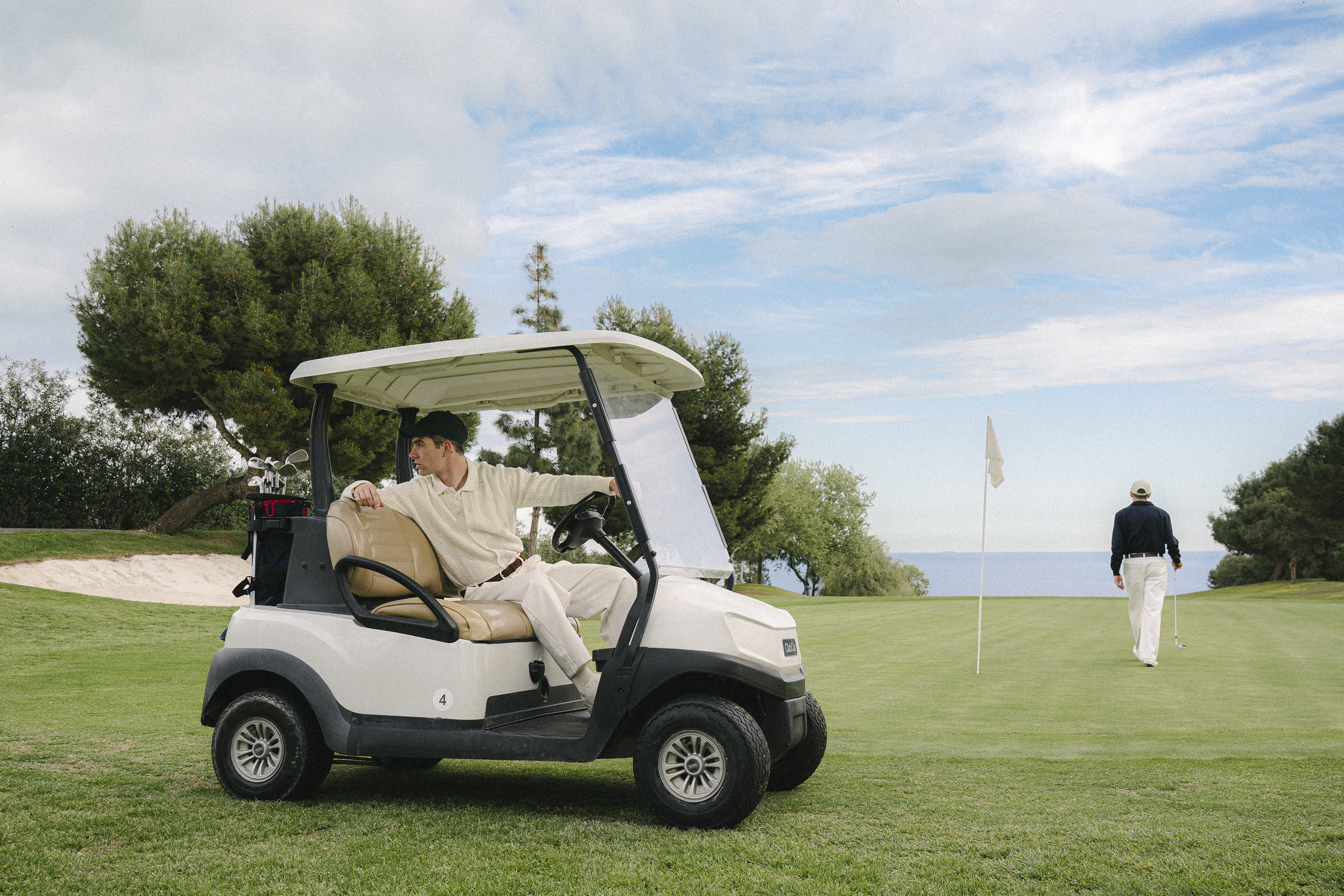 Golfer in golf cart on sunny green course. Man walks towards flag with ocean.