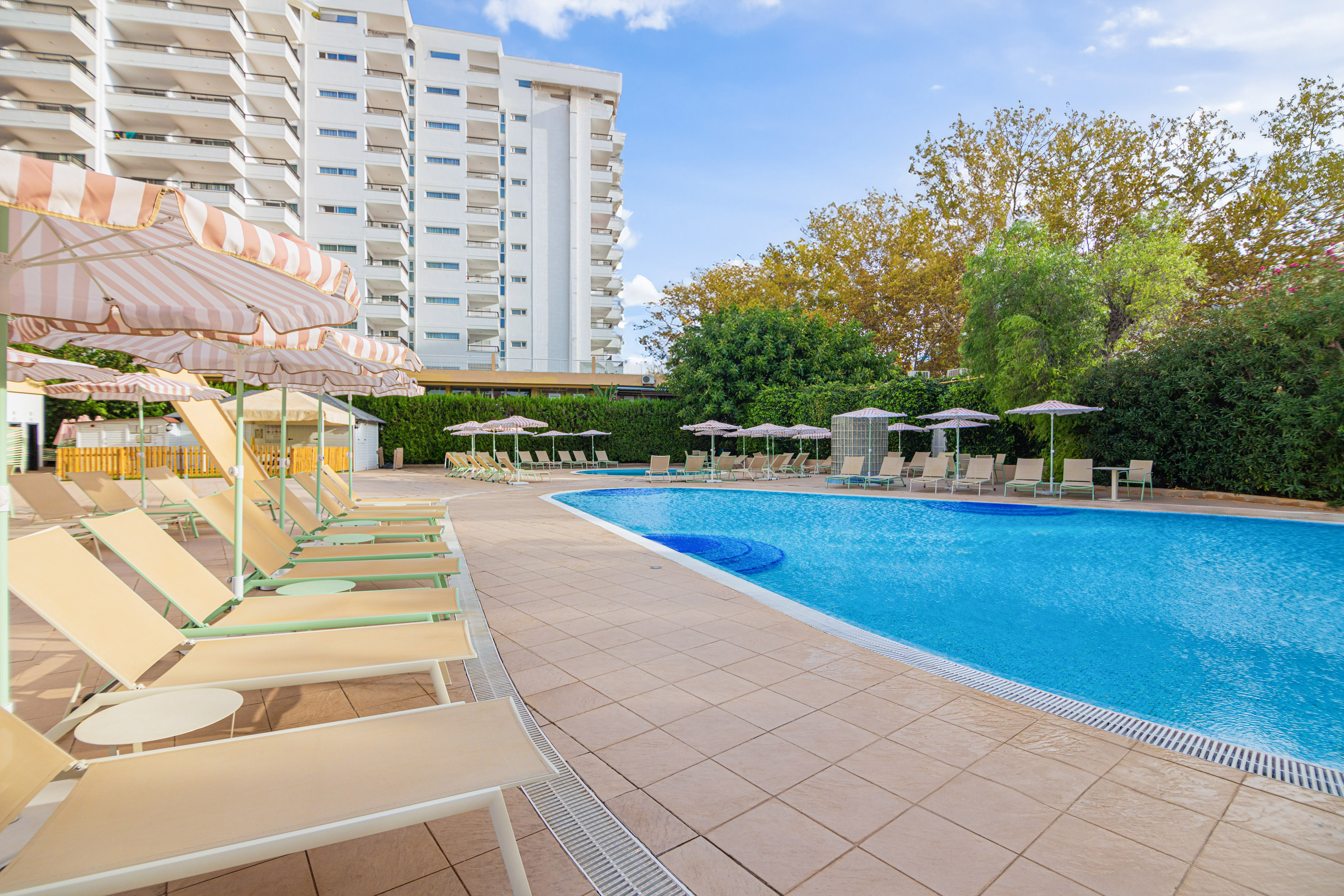 Sunny hotel pool with sun loungers, striped umbrellas, and a resort building.