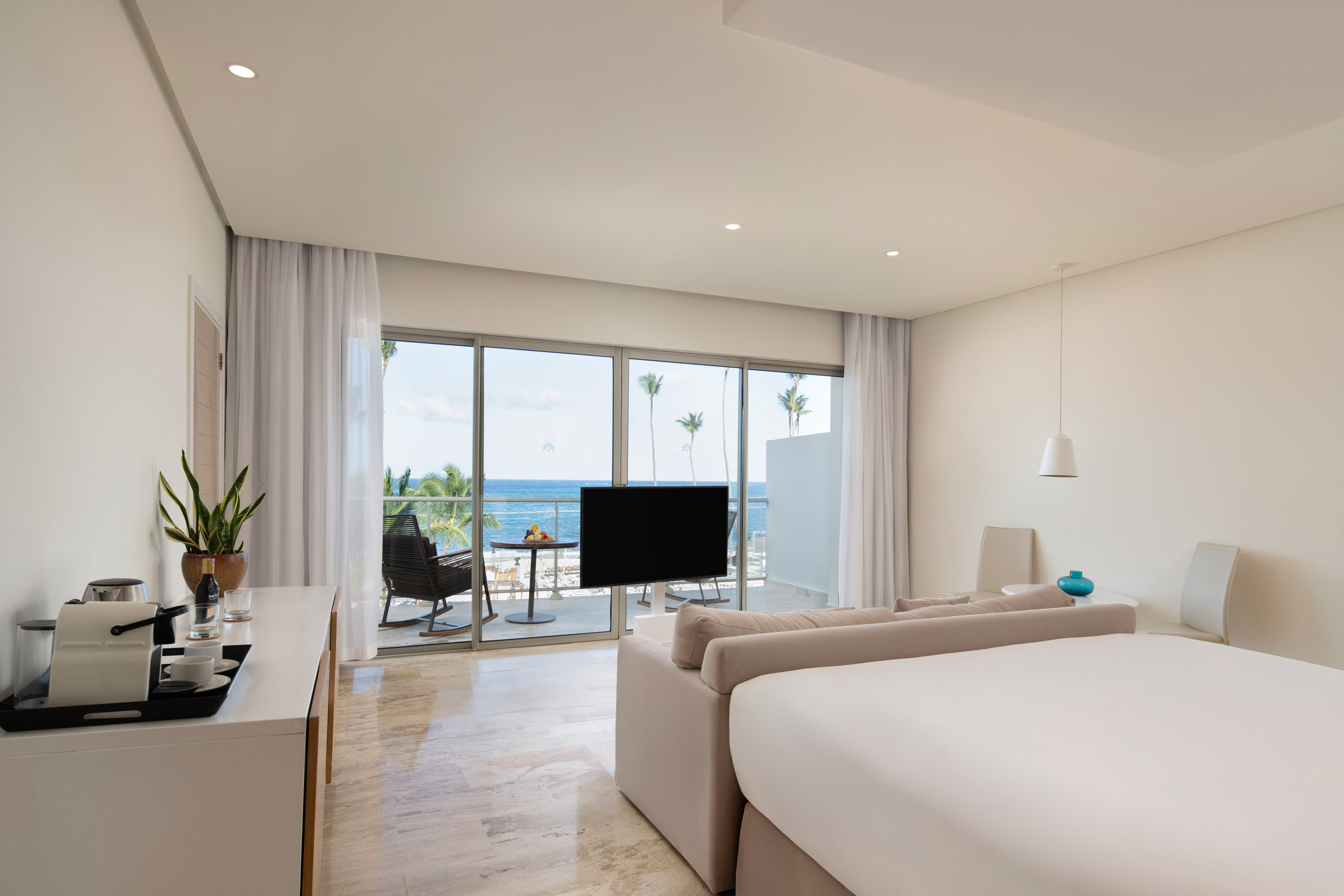 Modern ocean view hotel suite with balcony, king bed, and tropical palm trees.