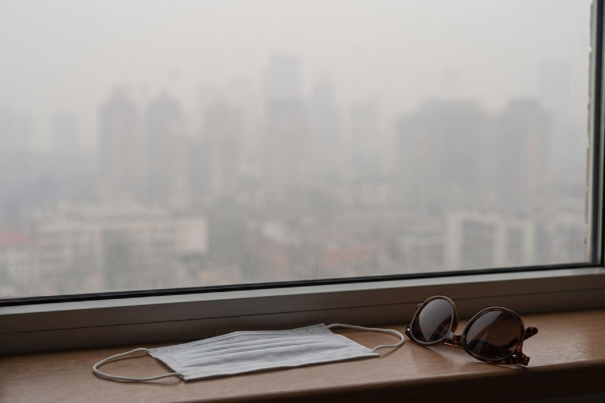 Mask and sunglasses on window sill, city view hazy from air pollution.
