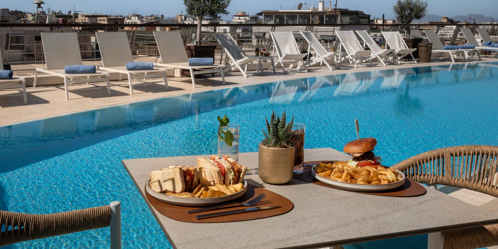 Rooftop pool with poolside dining, burger, sandwich, fries, and city view.