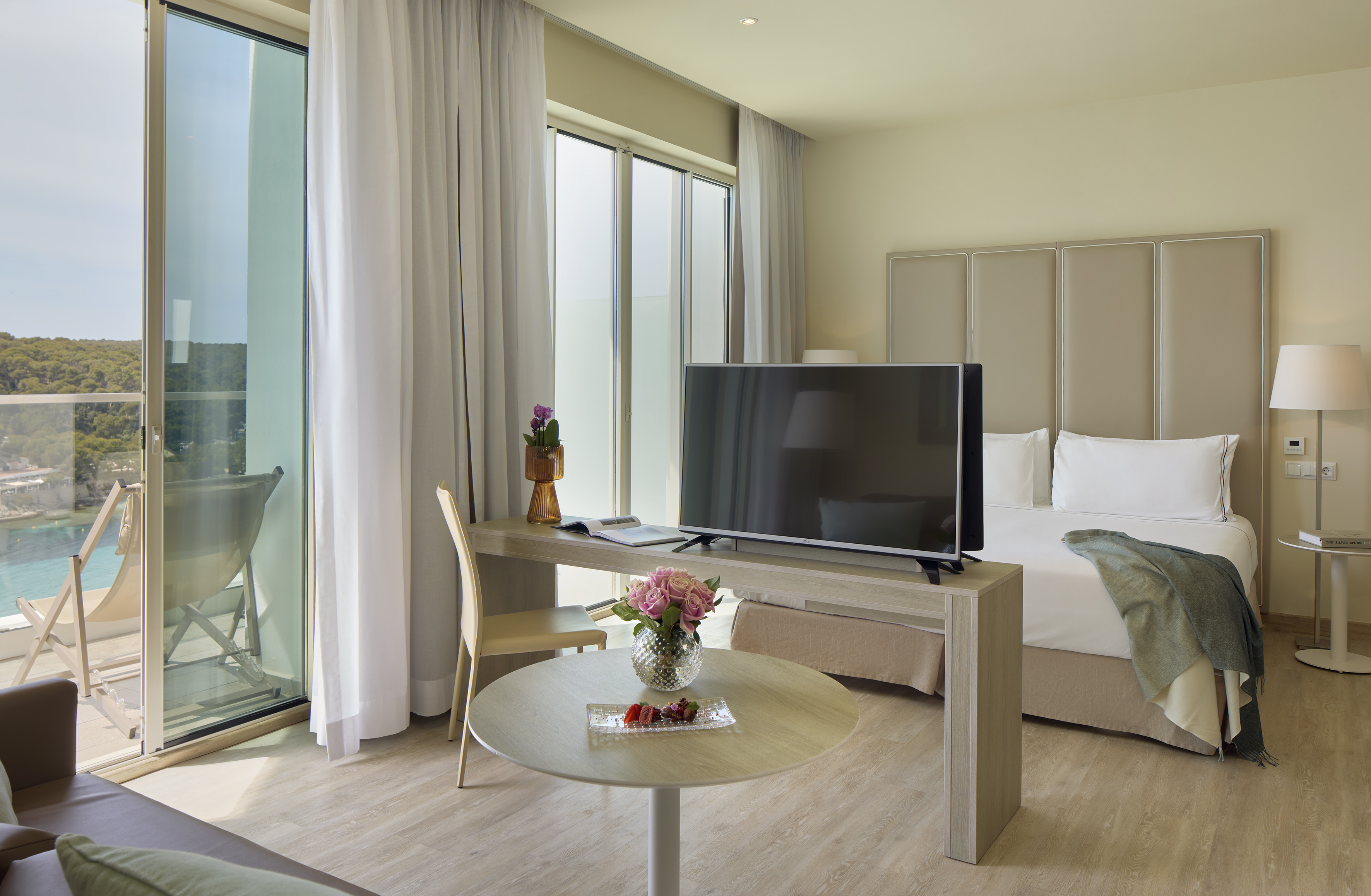 Modern hotel room with sea view balcony, bed, TV, and desk.