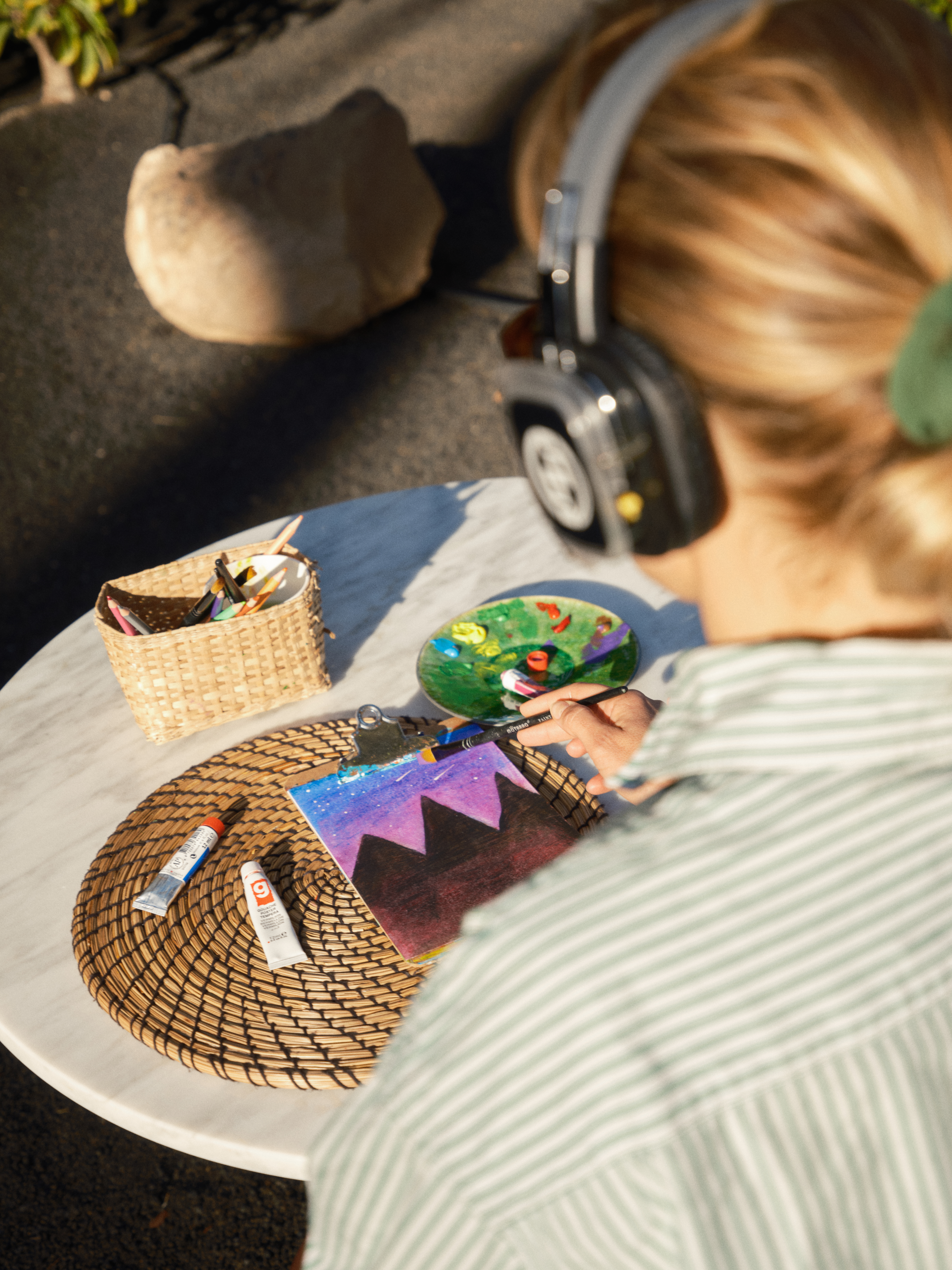 Person in headphones painting mountain night sky art on a clipboard outdoors.
