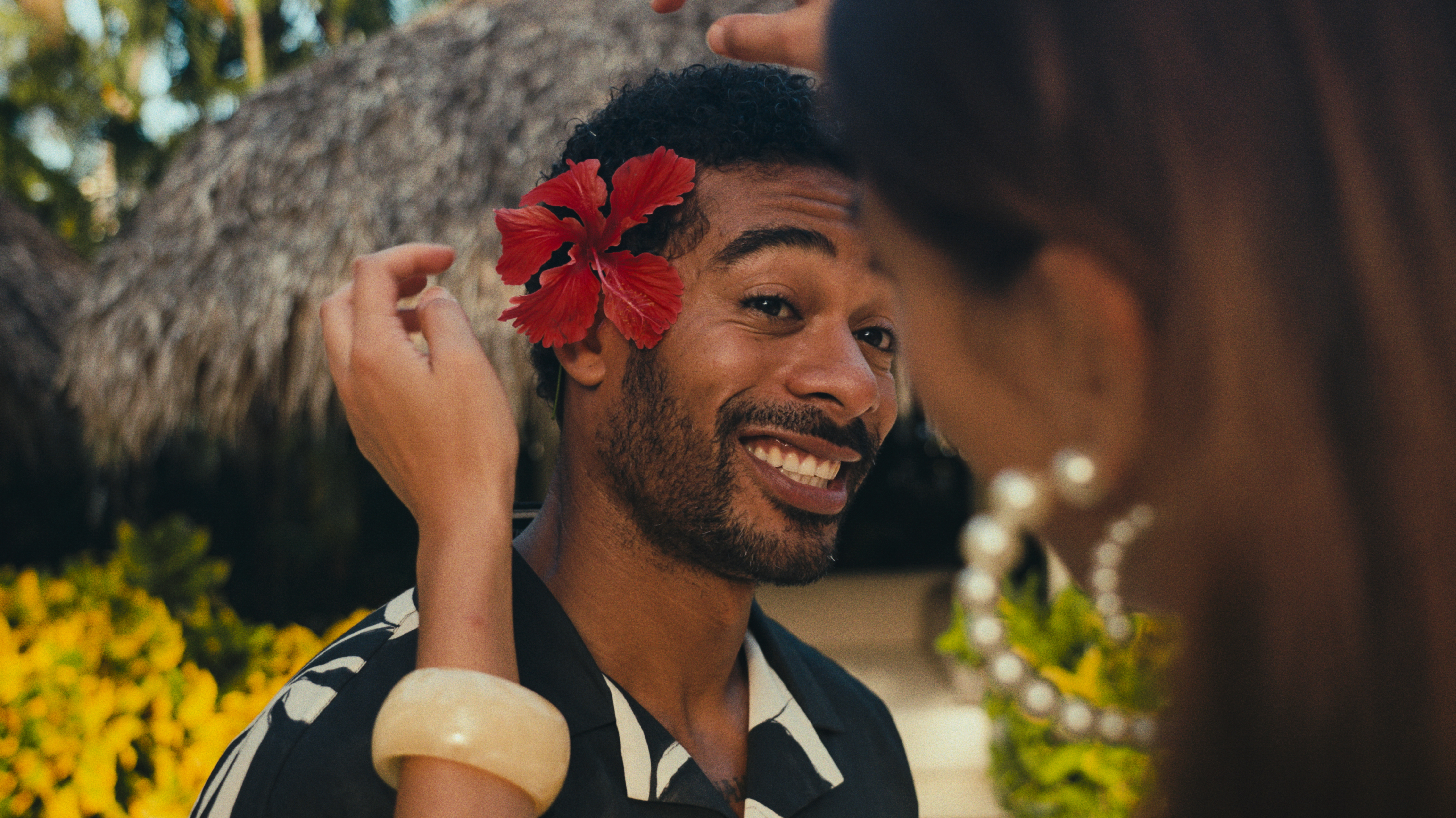 Woman placing red hibiscus flower behind smiling man's ear. Tropical setting.