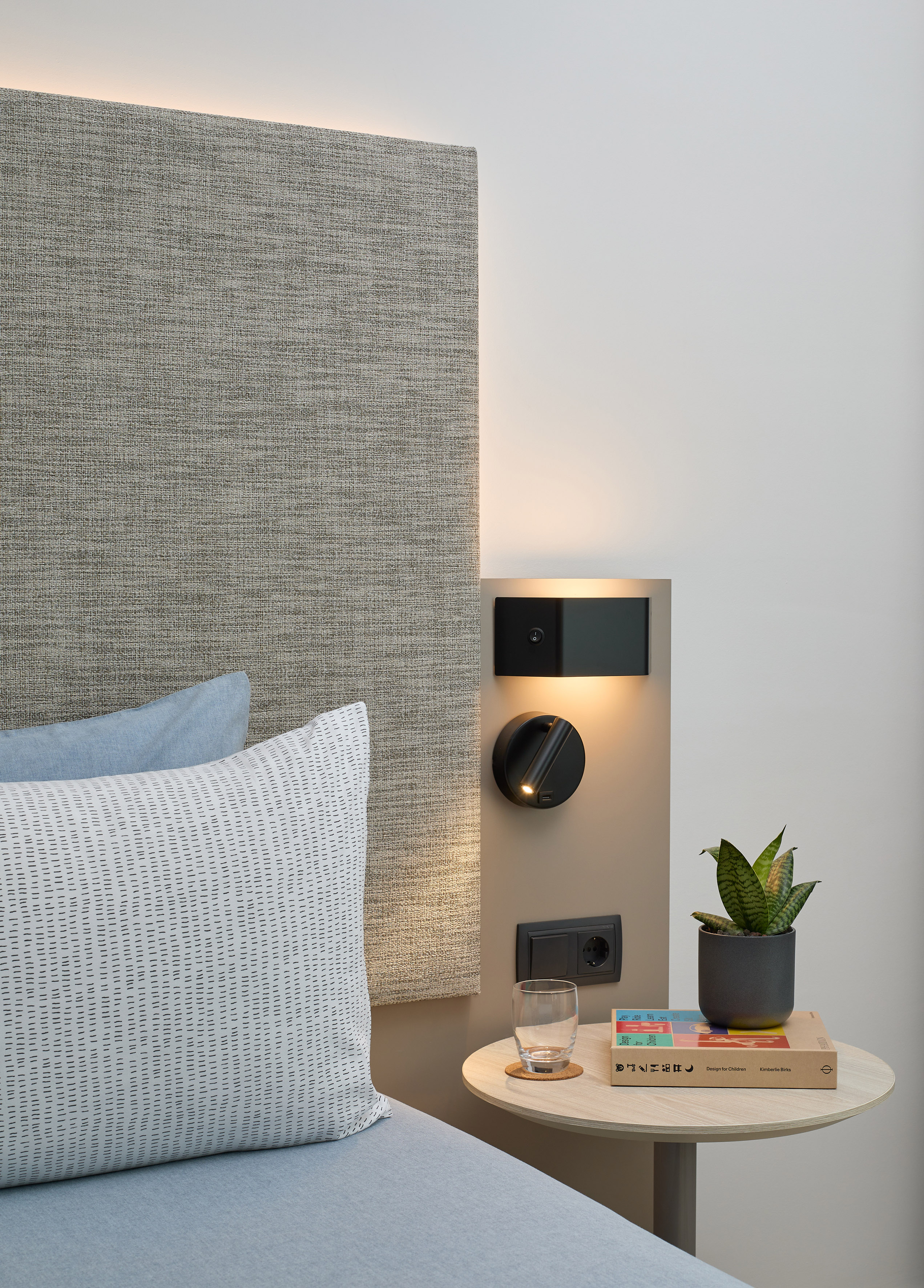 Modern hotel bedroom nightstand with lamp, plant, book, textured headboard.
