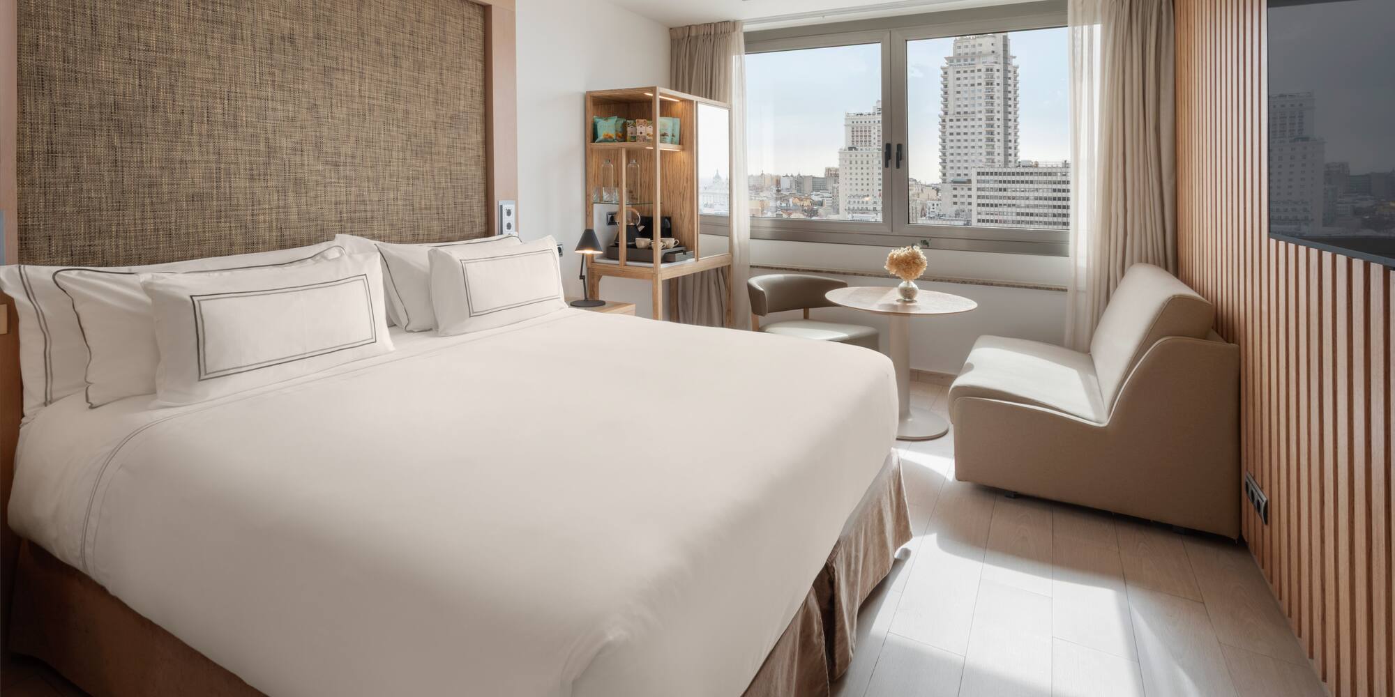 Modern hotel room with king bed, city view, and light wooden decor.