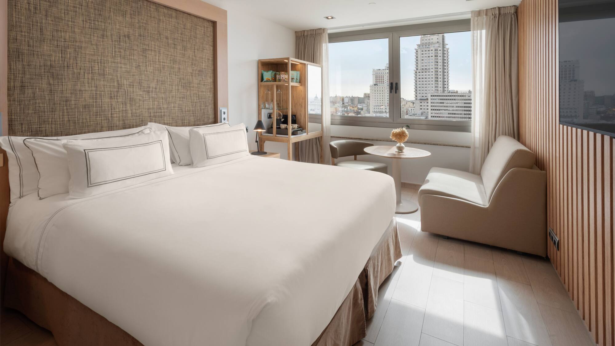 Modern hotel room with king bed, city view, and light wooden decor.