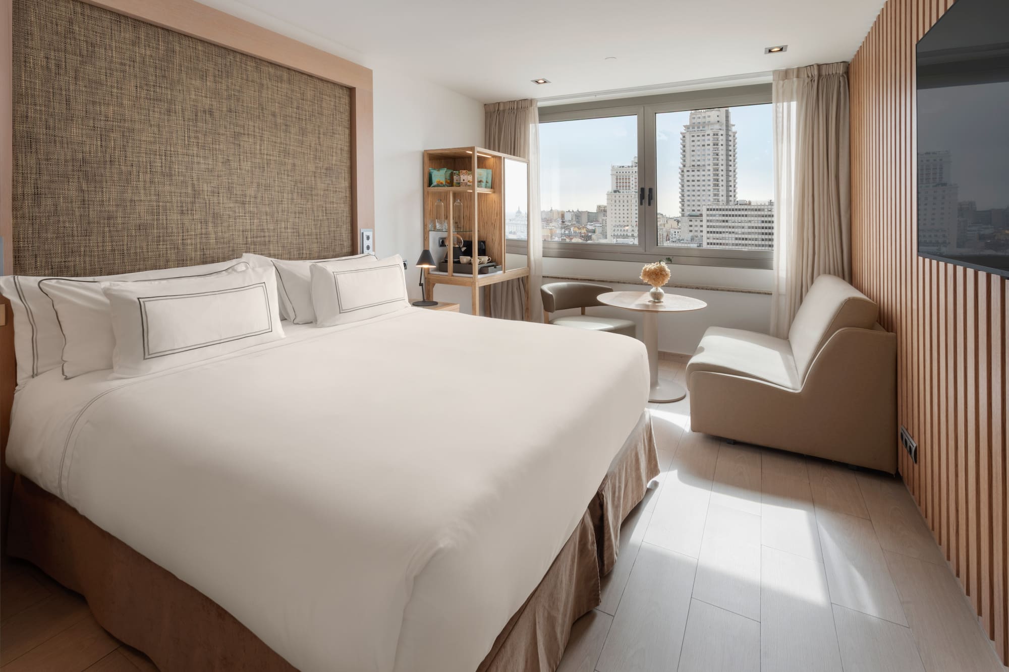 Modern hotel room with king bed, city view, and light wooden decor.