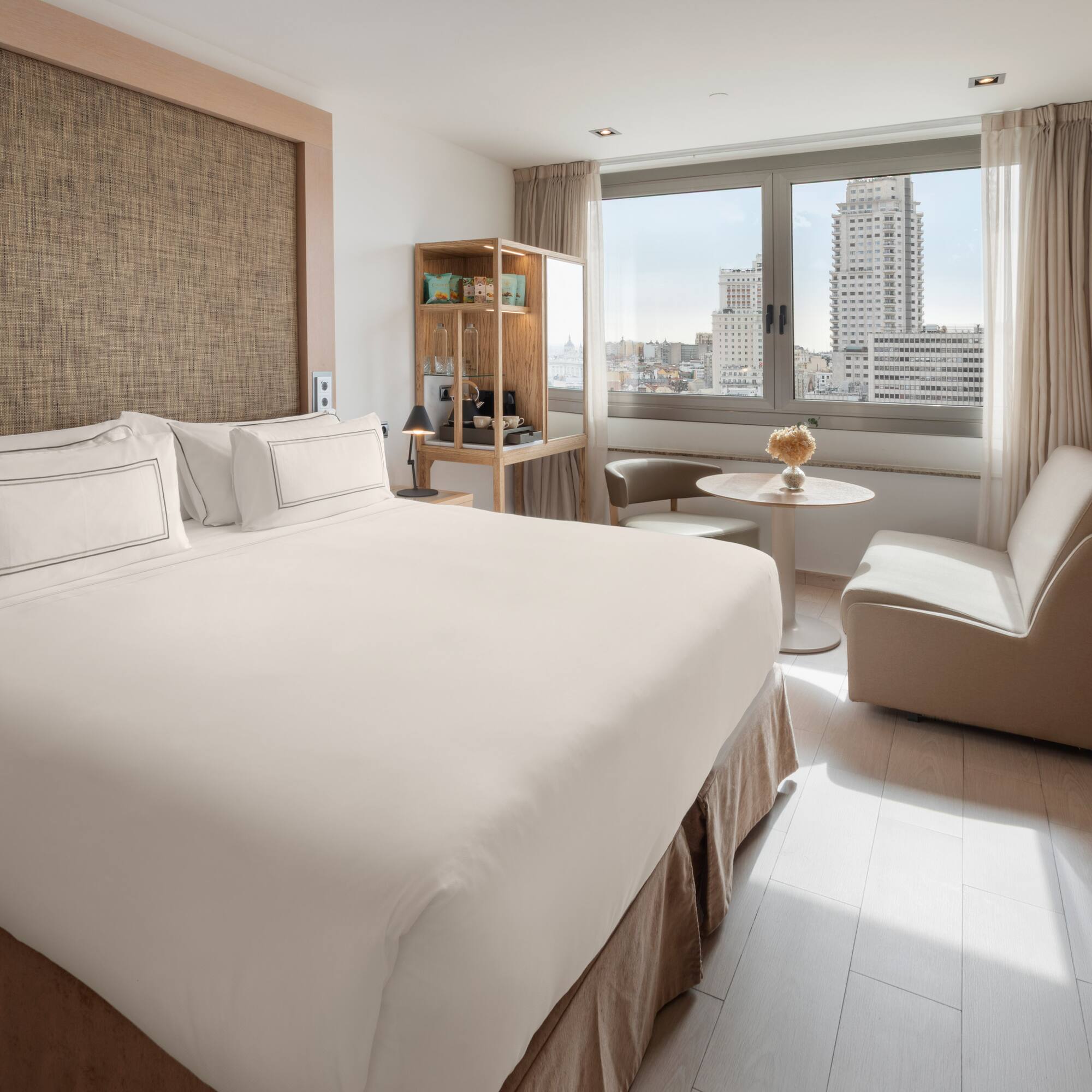Modern hotel room with king bed, city view, and light wooden decor.