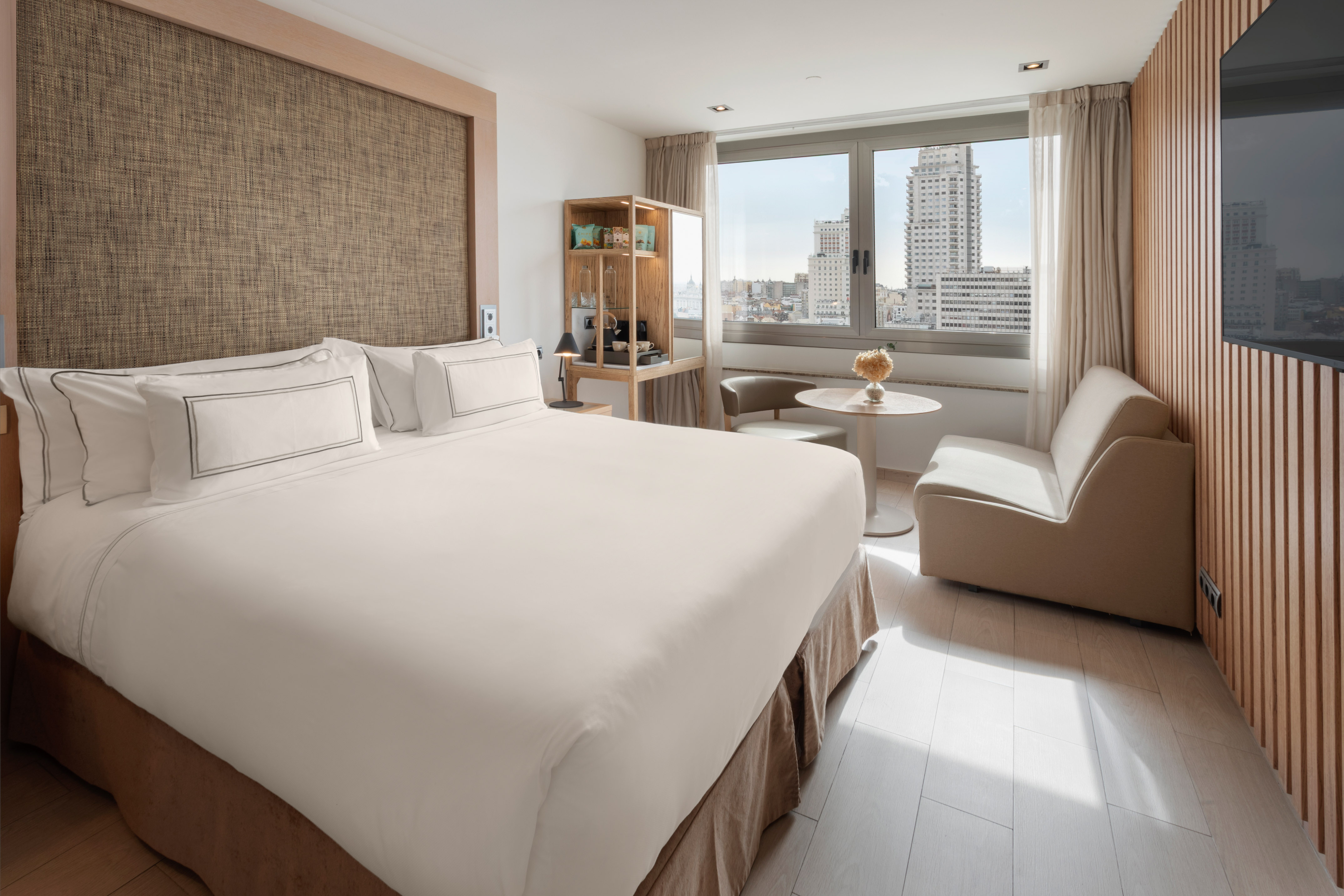 Modern hotel room with king bed, city view, and light wooden decor.