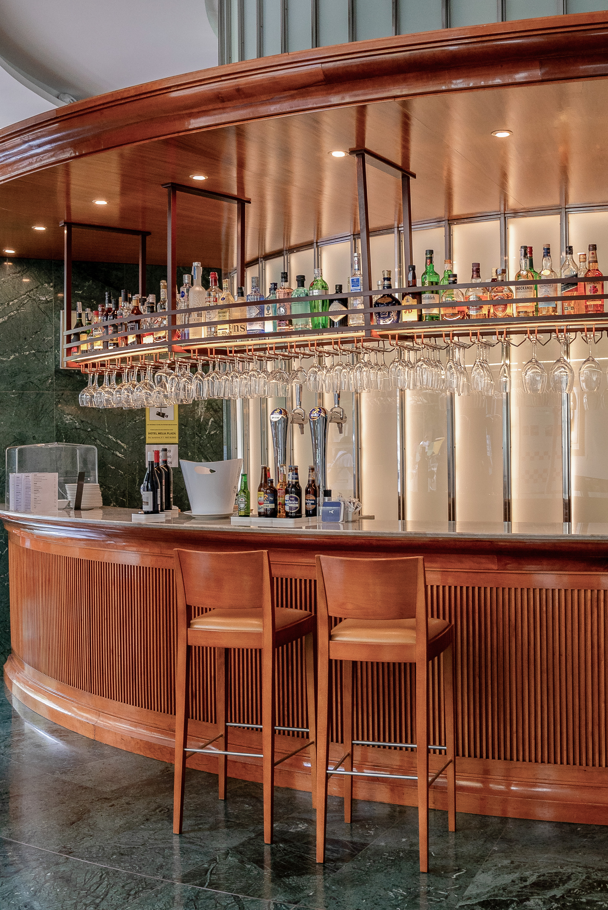 Wooden hotel bar with liquor bottles, hanging glasses, and two bar stools.