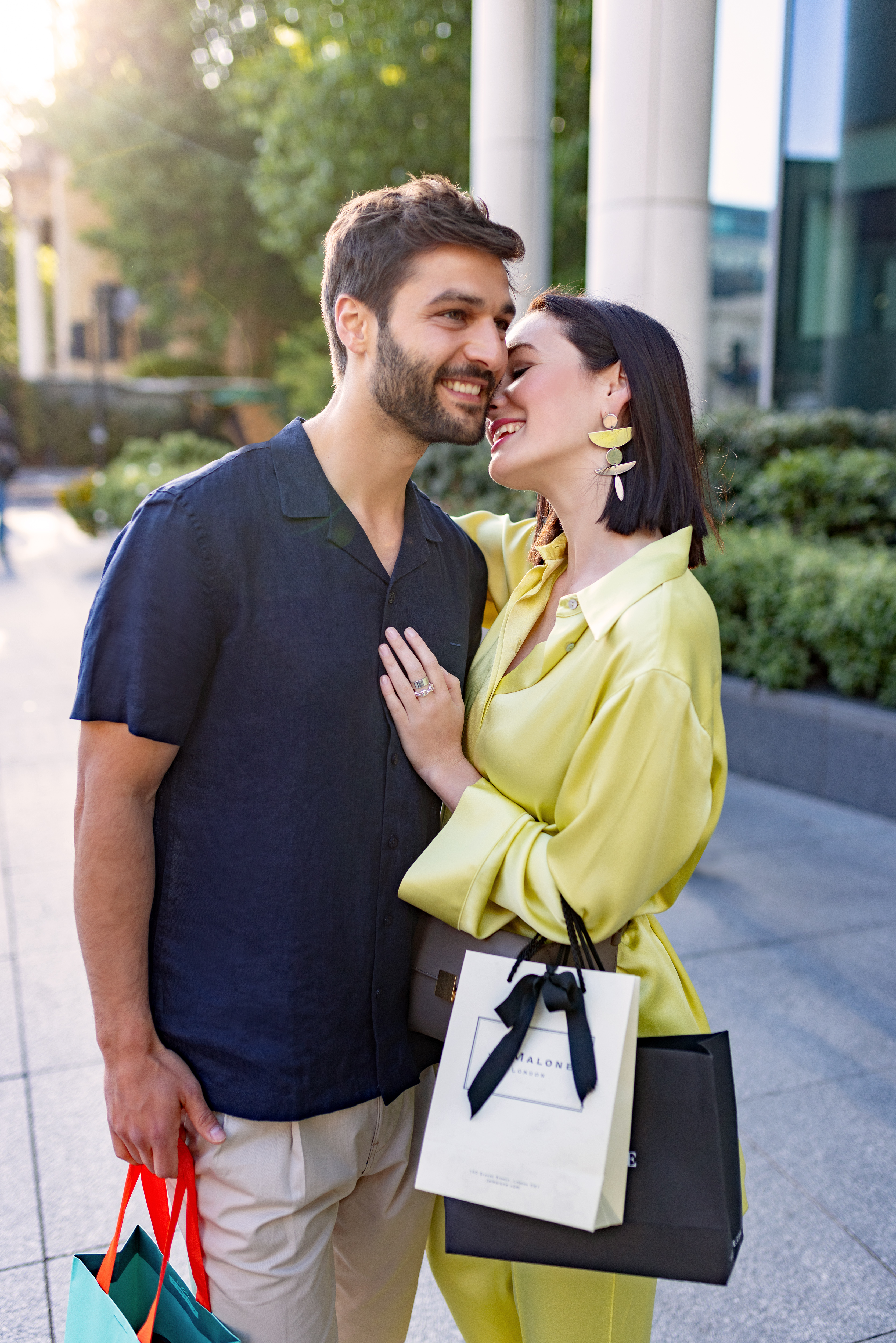 Smiling couple embracing, holding Jo Malone and Tiffany shopping bags, urban street.