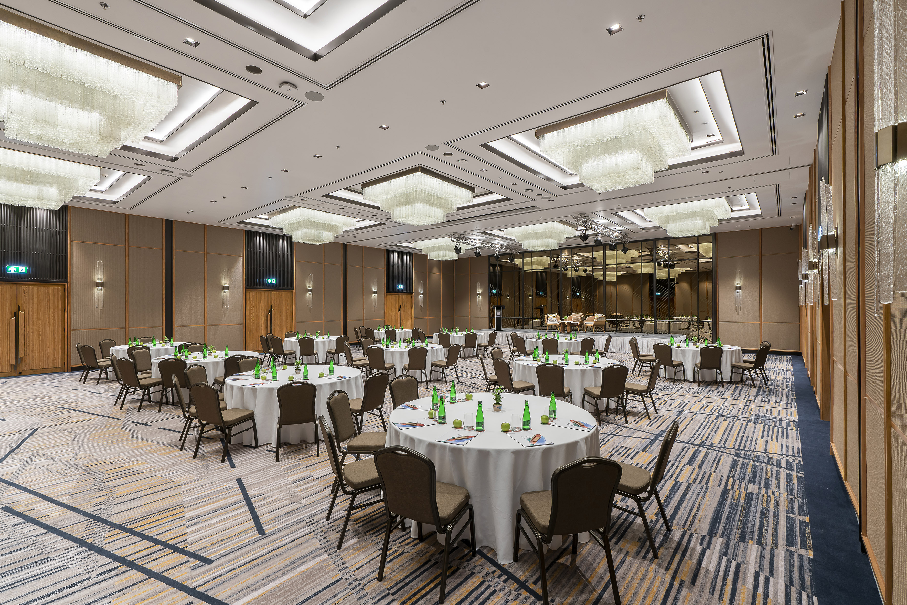 Elegant banquet hall setup with round tables, chairs, chandeliers, and stage.