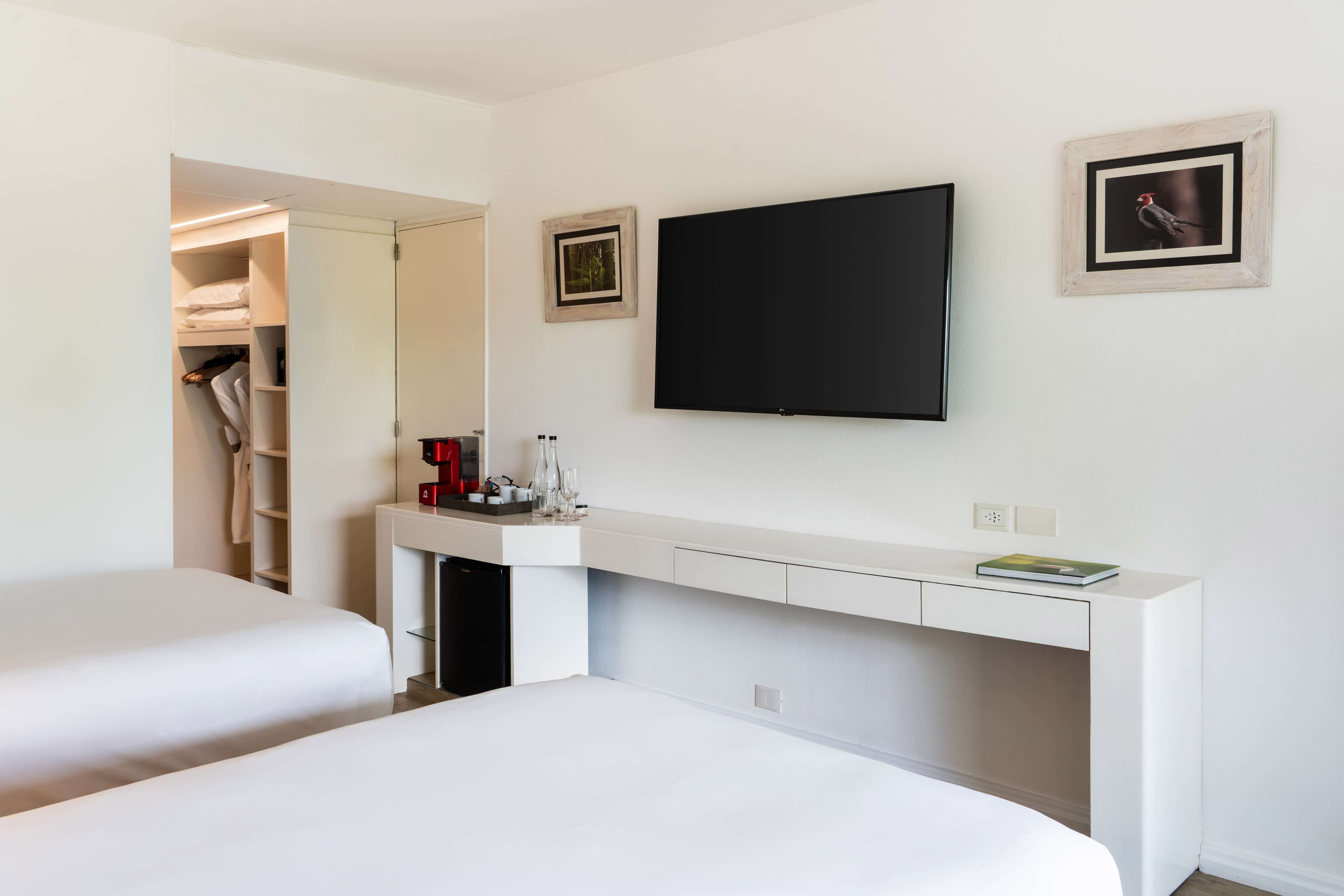 Modern hotel room with two white beds, wall TV, coffee station, and open closet.