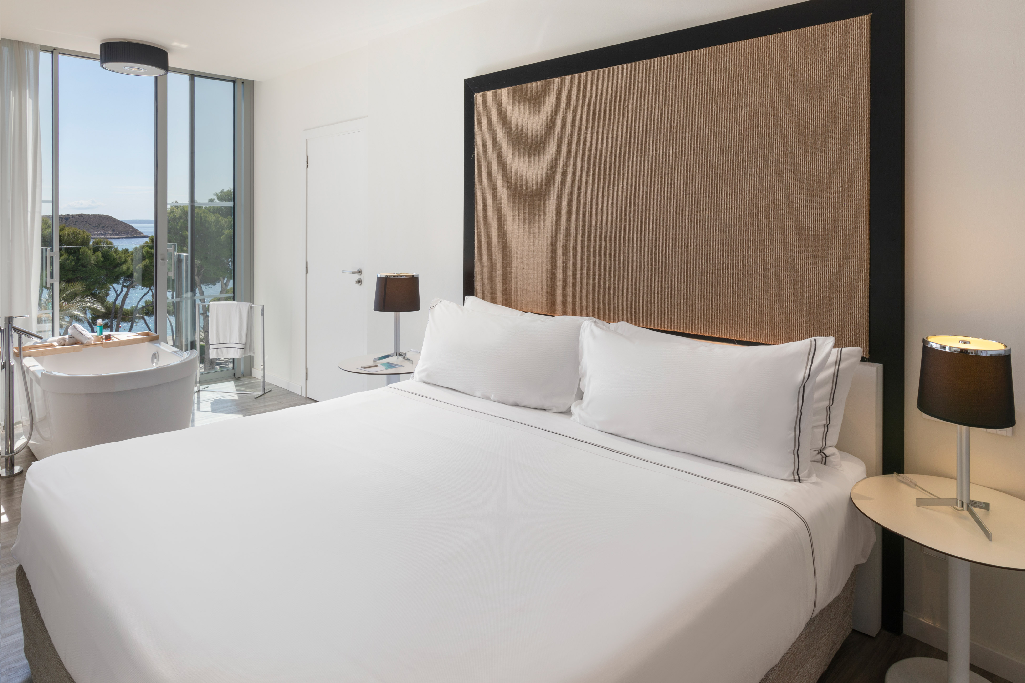 Modern luxury hotel bedroom with white bed, sea view, and freestanding tub.