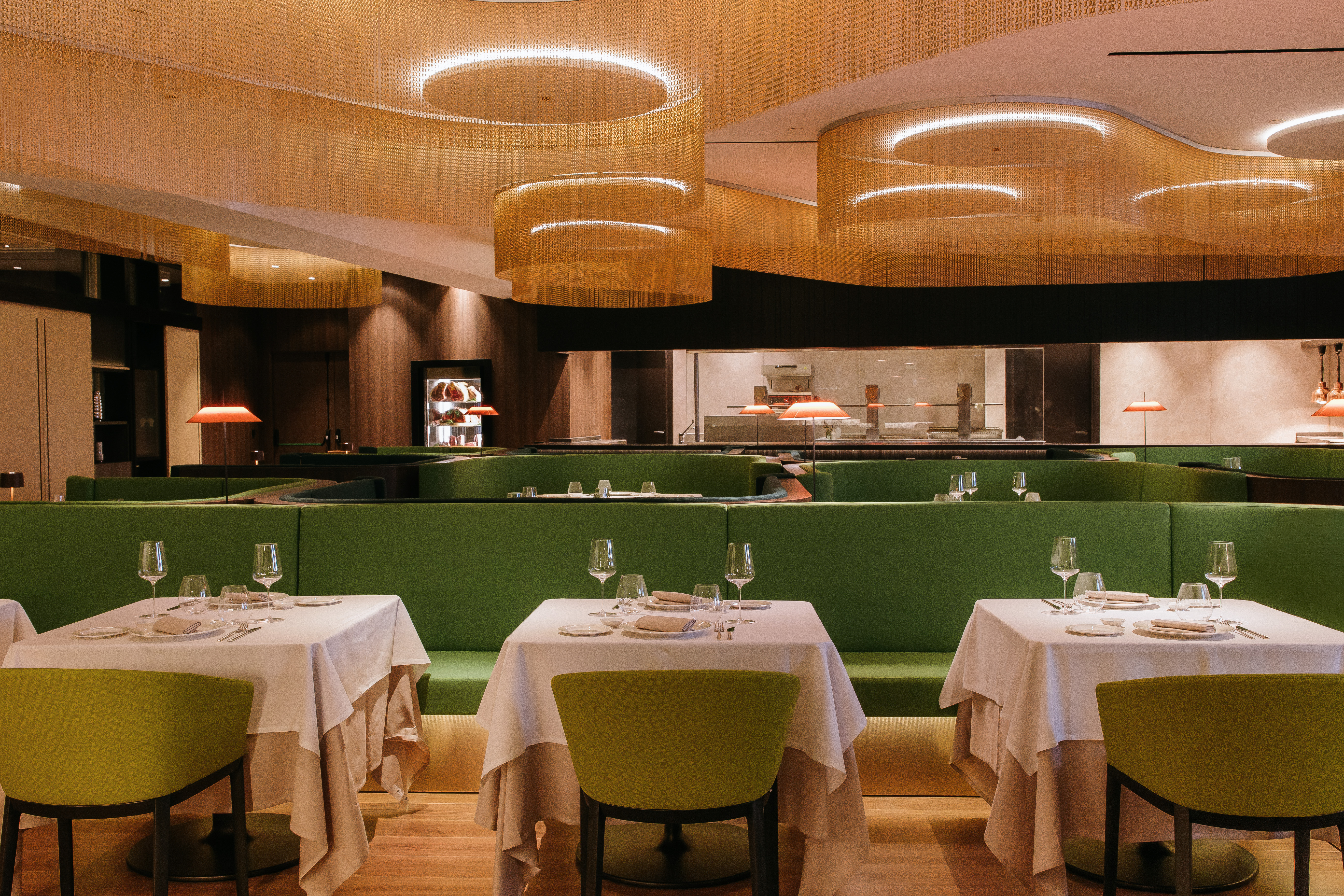 Modern restaurant with green booths, white tables, and golden chain ceiling decor.
