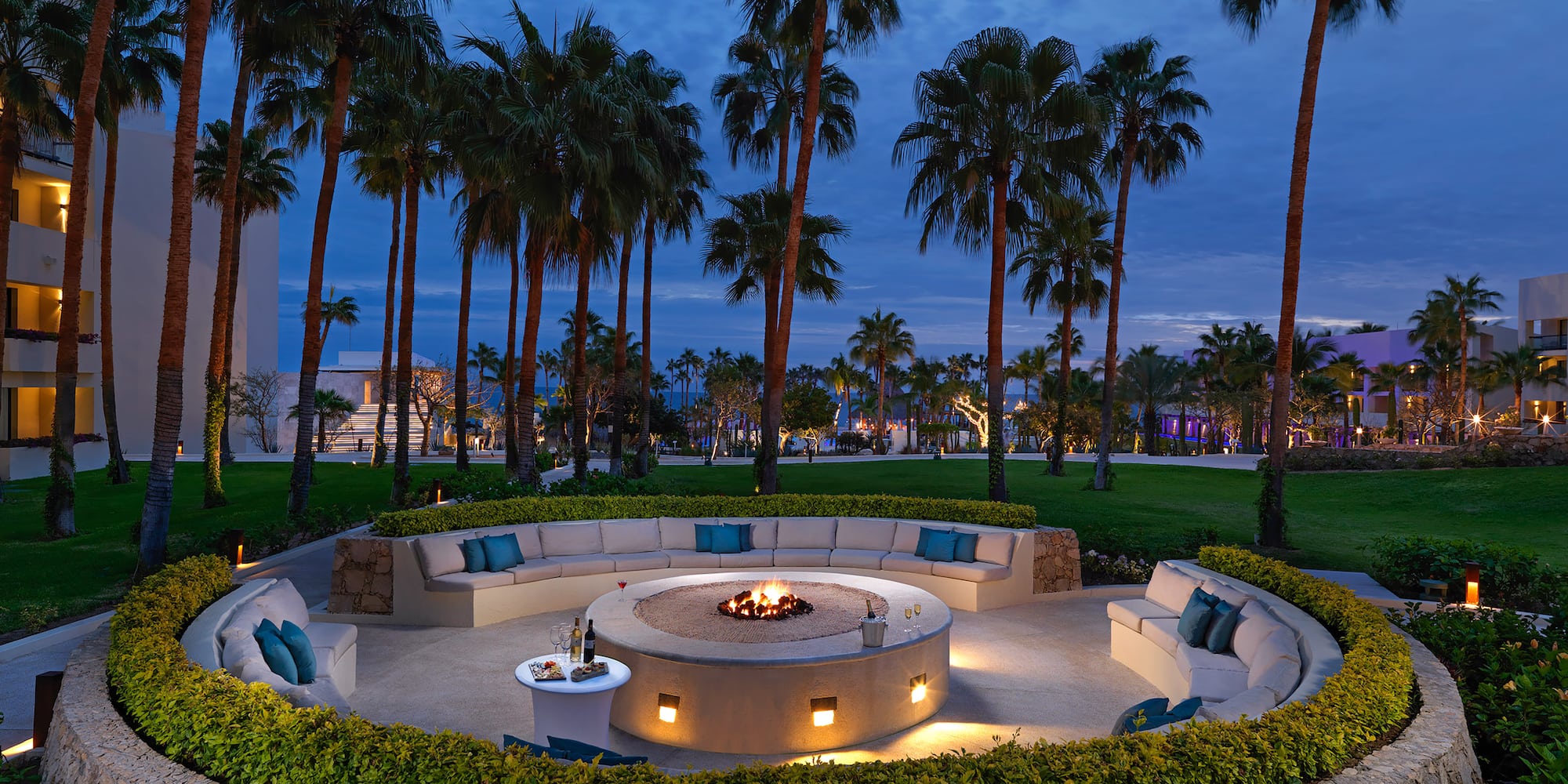 Outdoor resort fire pit lounge with curved seating, palm trees, and evening.