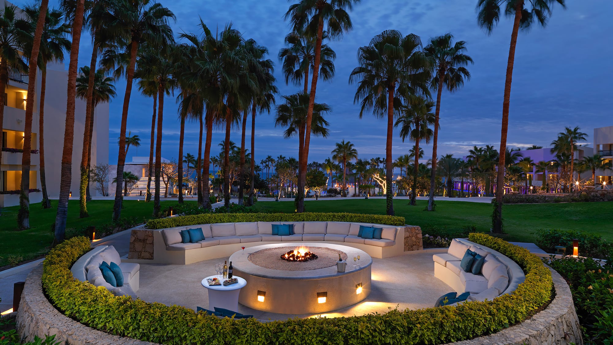 Outdoor resort fire pit lounge with curved seating, palm trees, and evening.