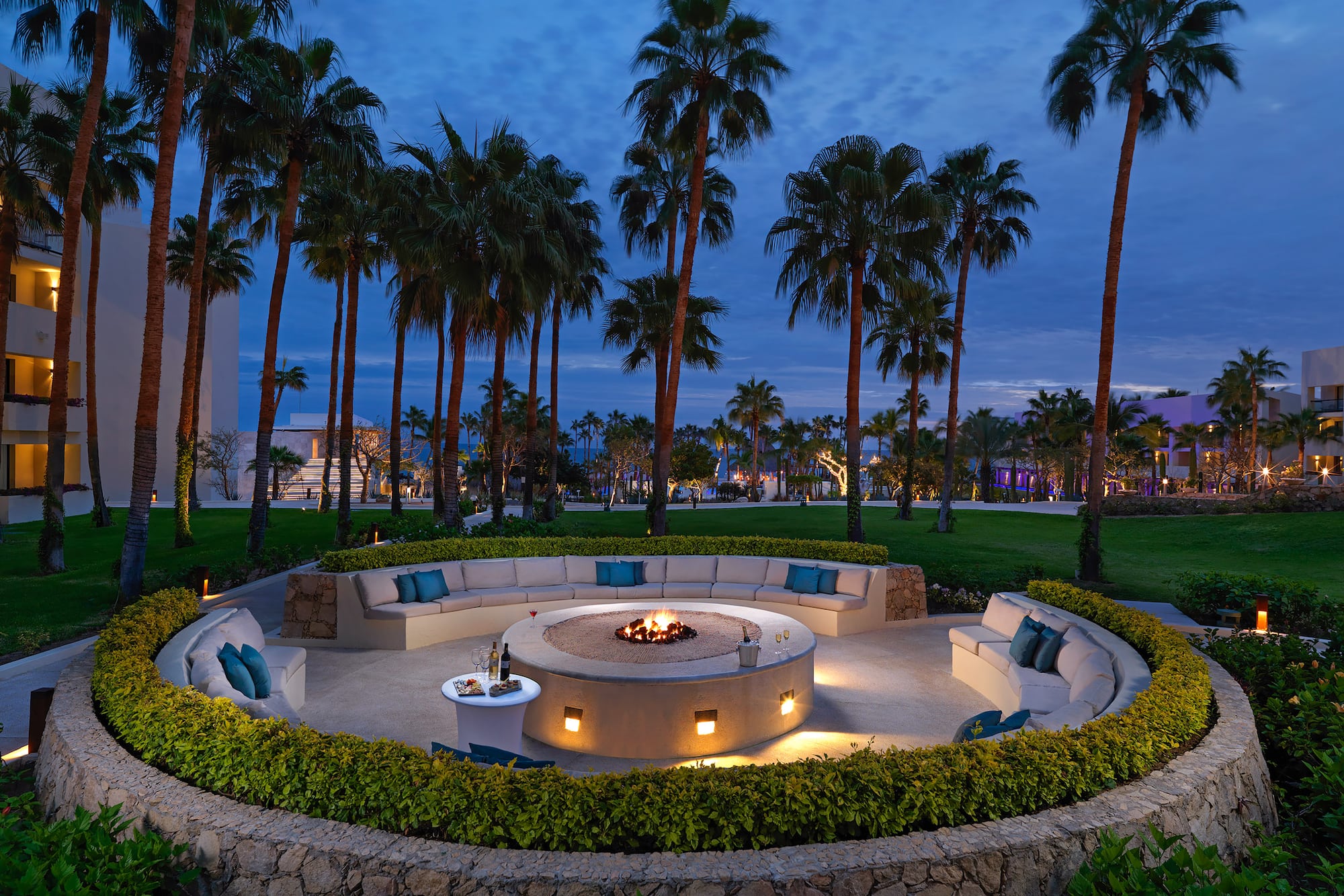 Outdoor resort fire pit lounge with curved seating, palm trees, and evening.