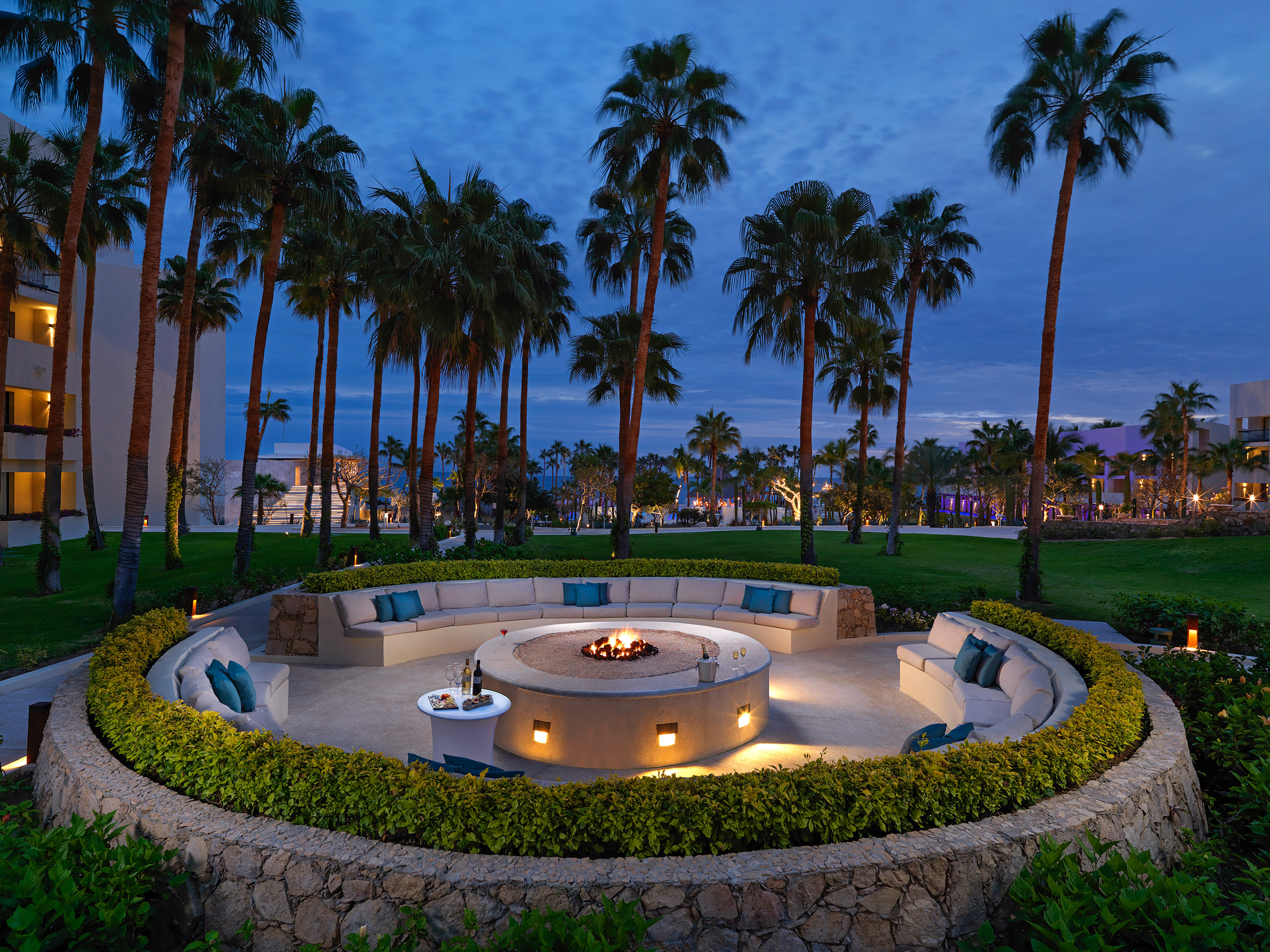 Outdoor resort fire pit lounge with curved seating, palm trees, and evening.