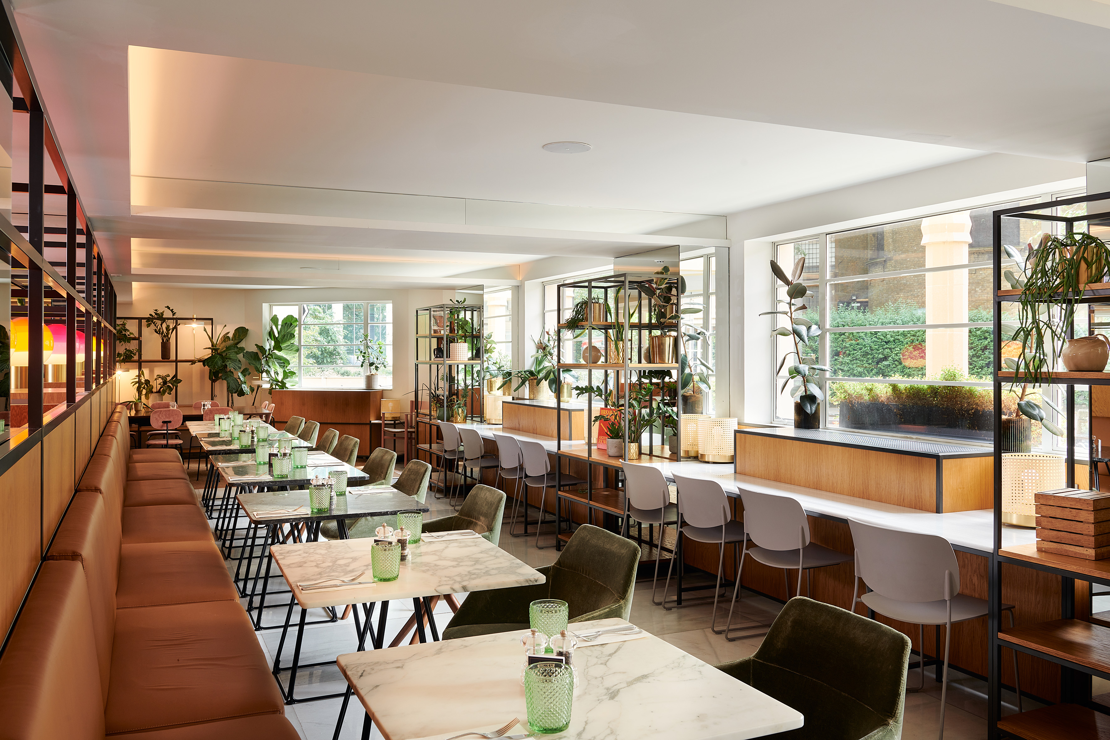 Modern restaurant interior with marble tables, banquette seating, and abundant plants.