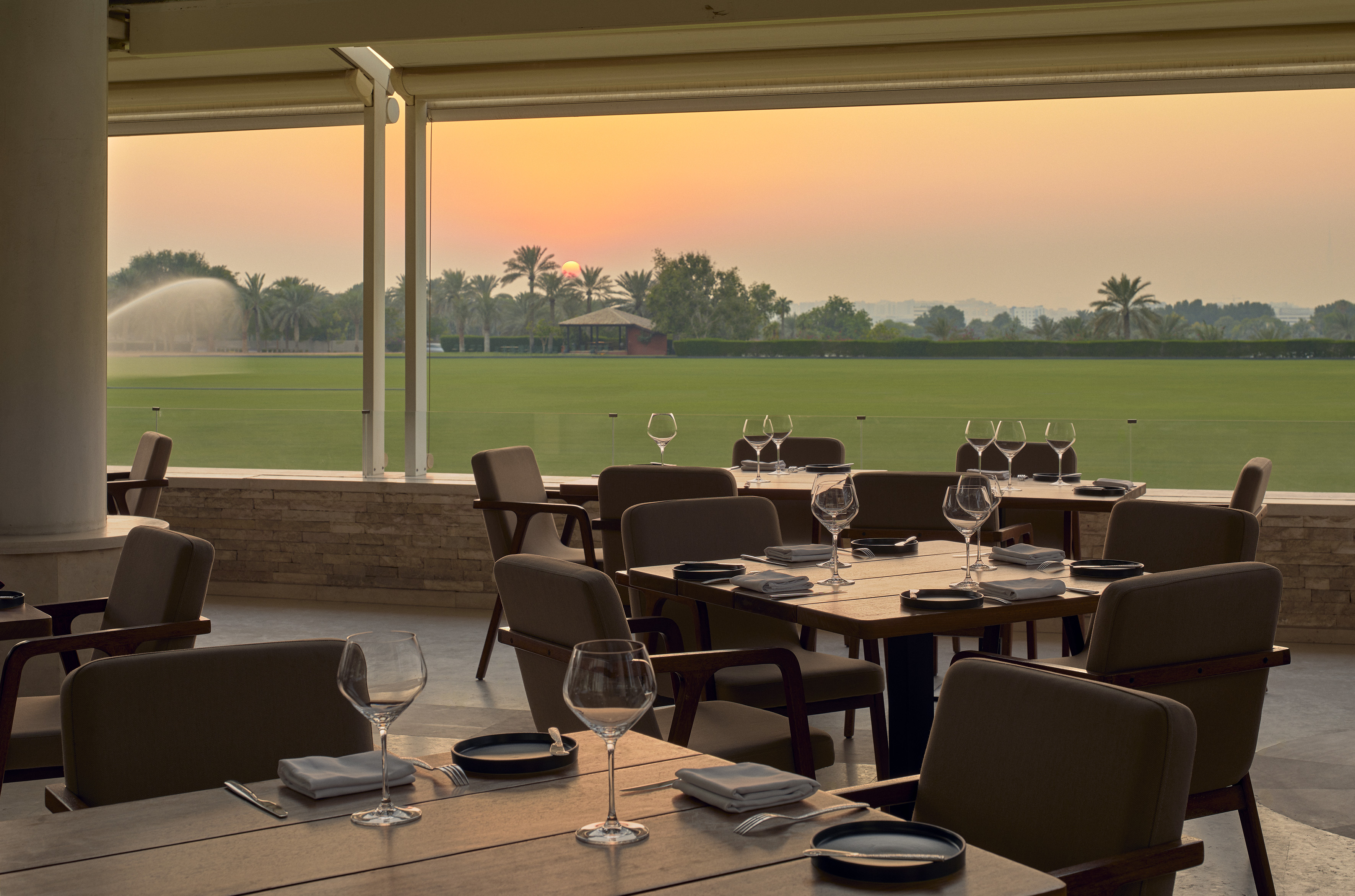 Outdoor restaurant patio with dining tables, sunset over green polo field.