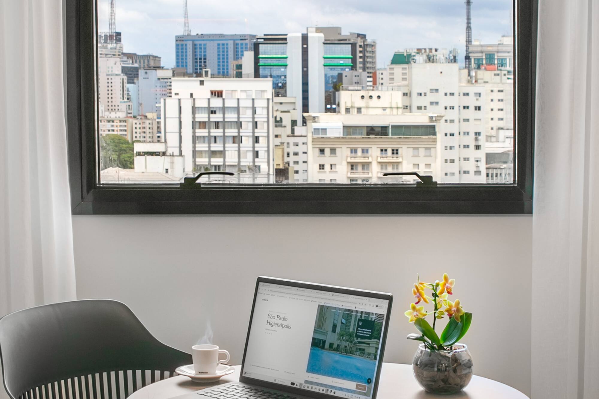 Work desk with laptop and coffee cup by window, overlooking city skyline.