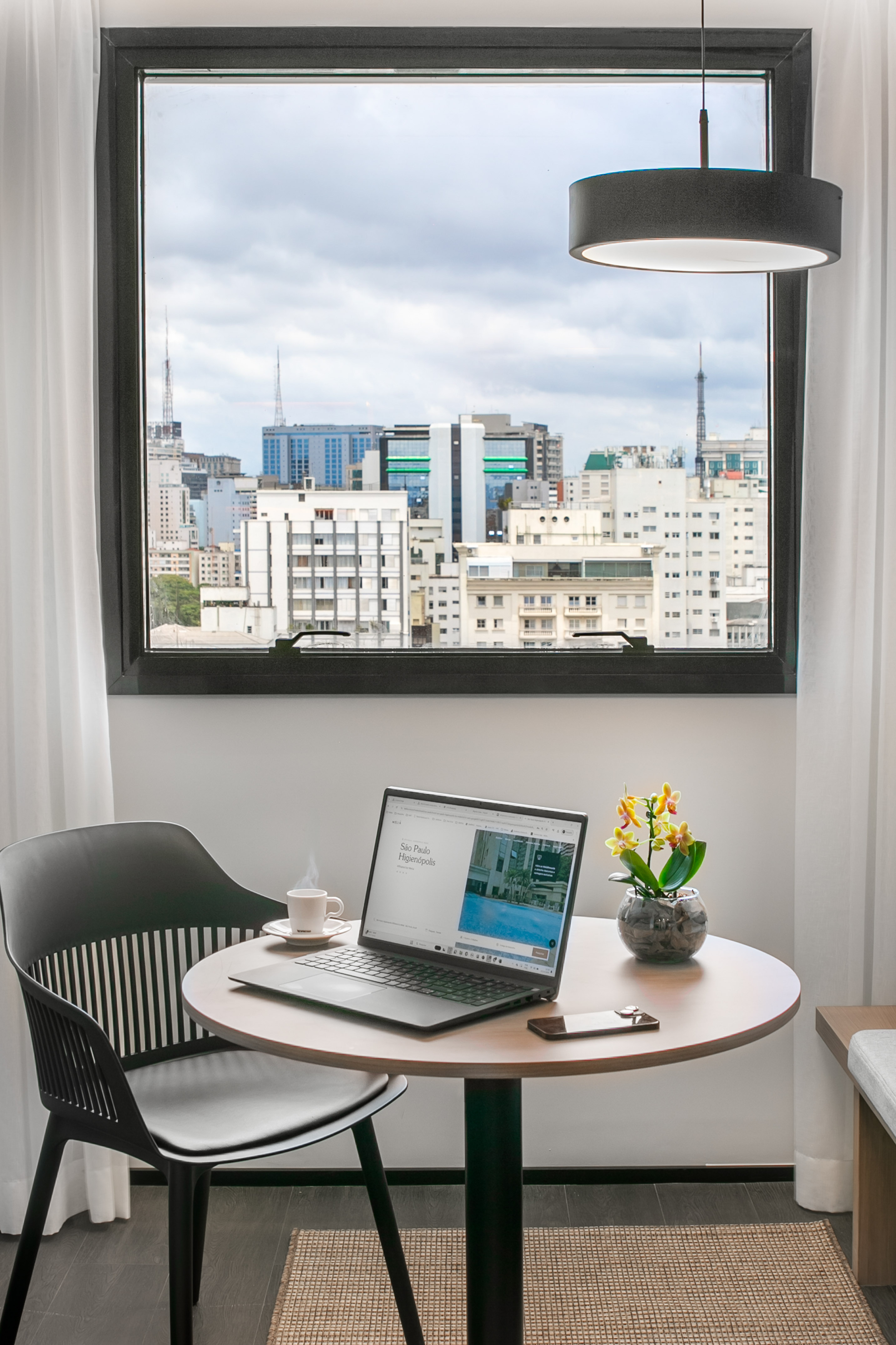 Work desk with laptop and coffee cup by window, overlooking city skyline.