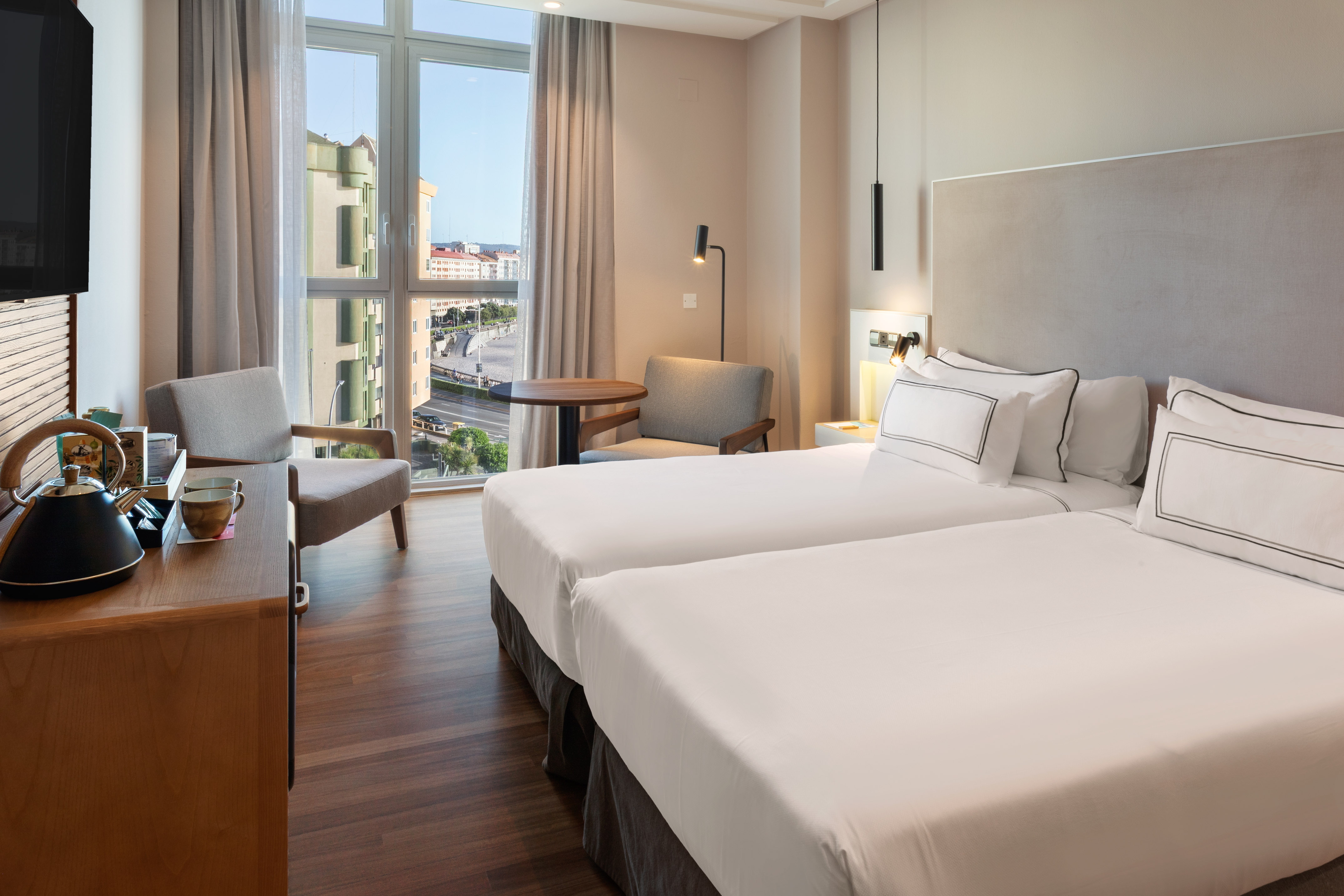 Modern hotel room with twin beds, city view, wood floor, and kettle station.