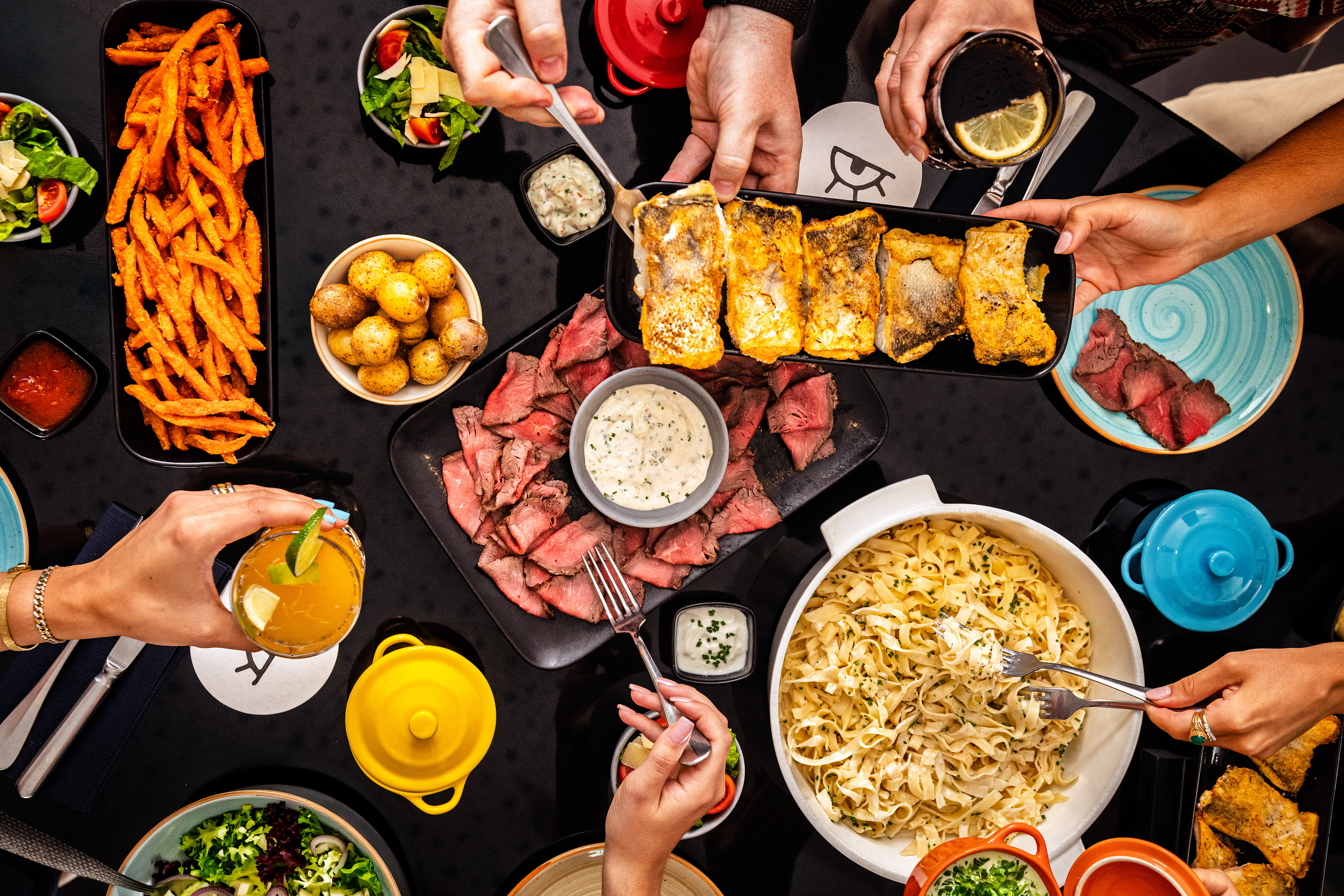 Hands sharing a vibrant dinner table: steak, fish, pasta, fries, salads, drinks.