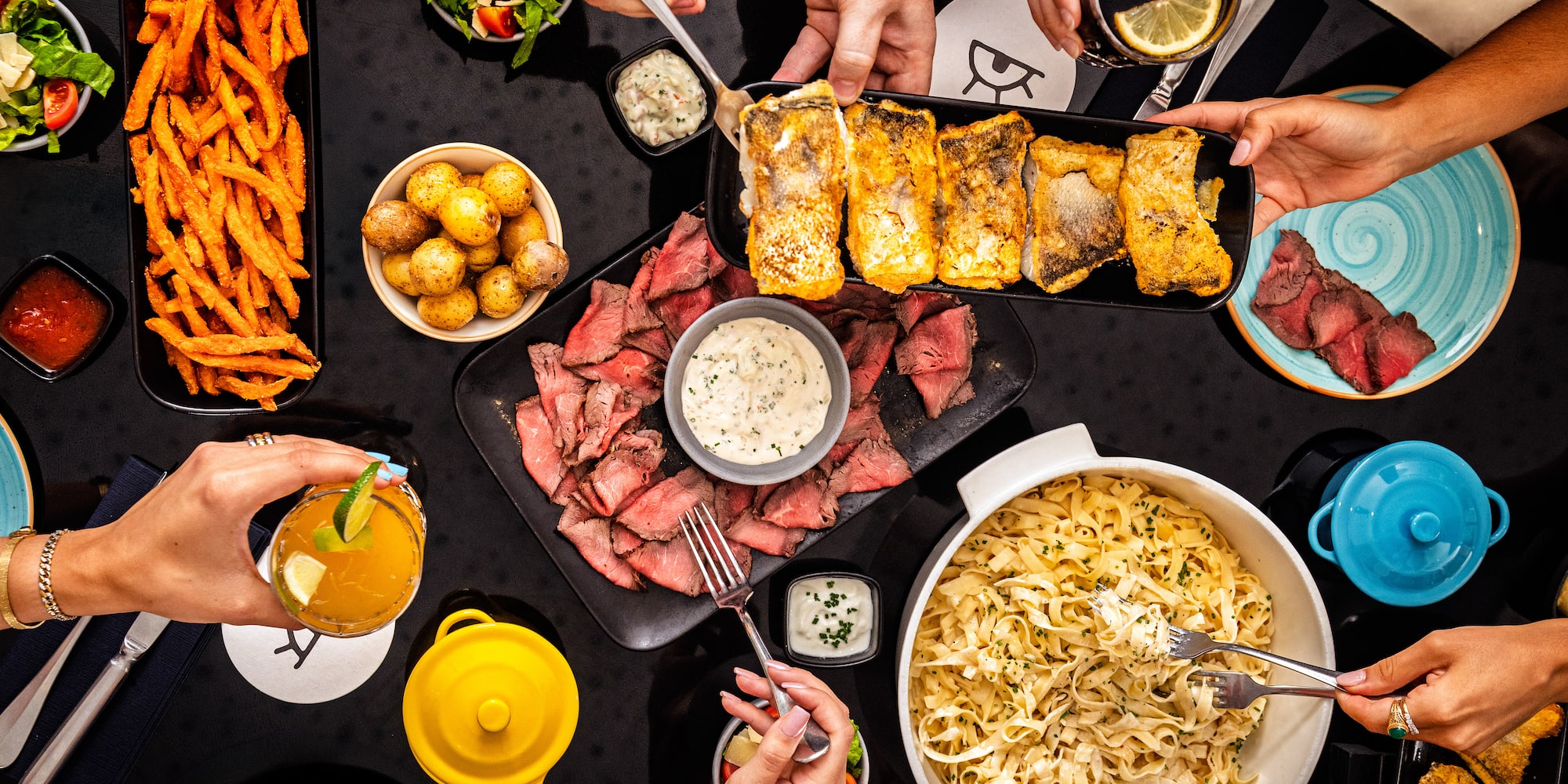 Hands sharing a vibrant dinner table: steak, fish, pasta, fries, salads, drinks.