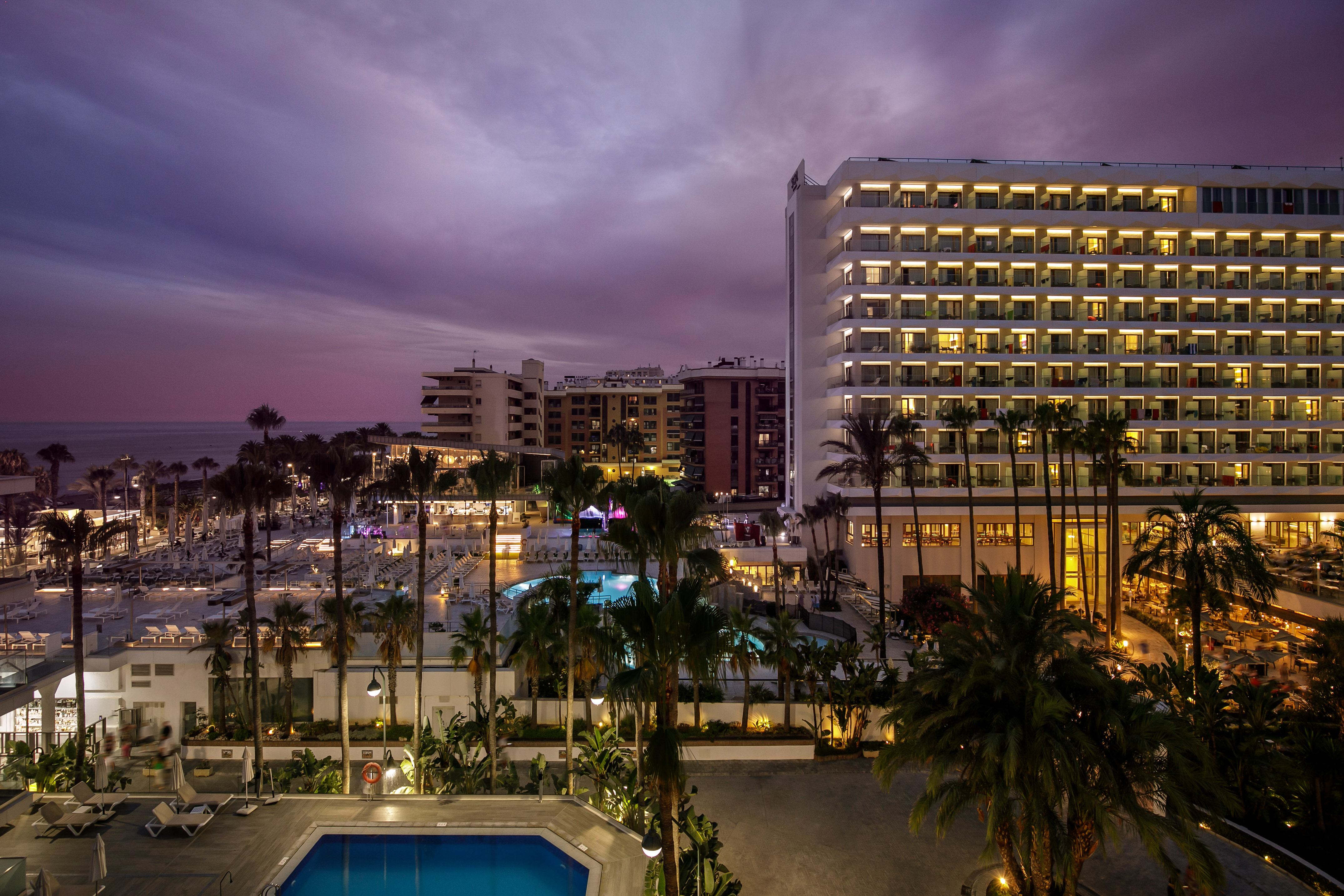 Beachfront hotel resort at dusk with illuminated pools, palm trees, and ocean.