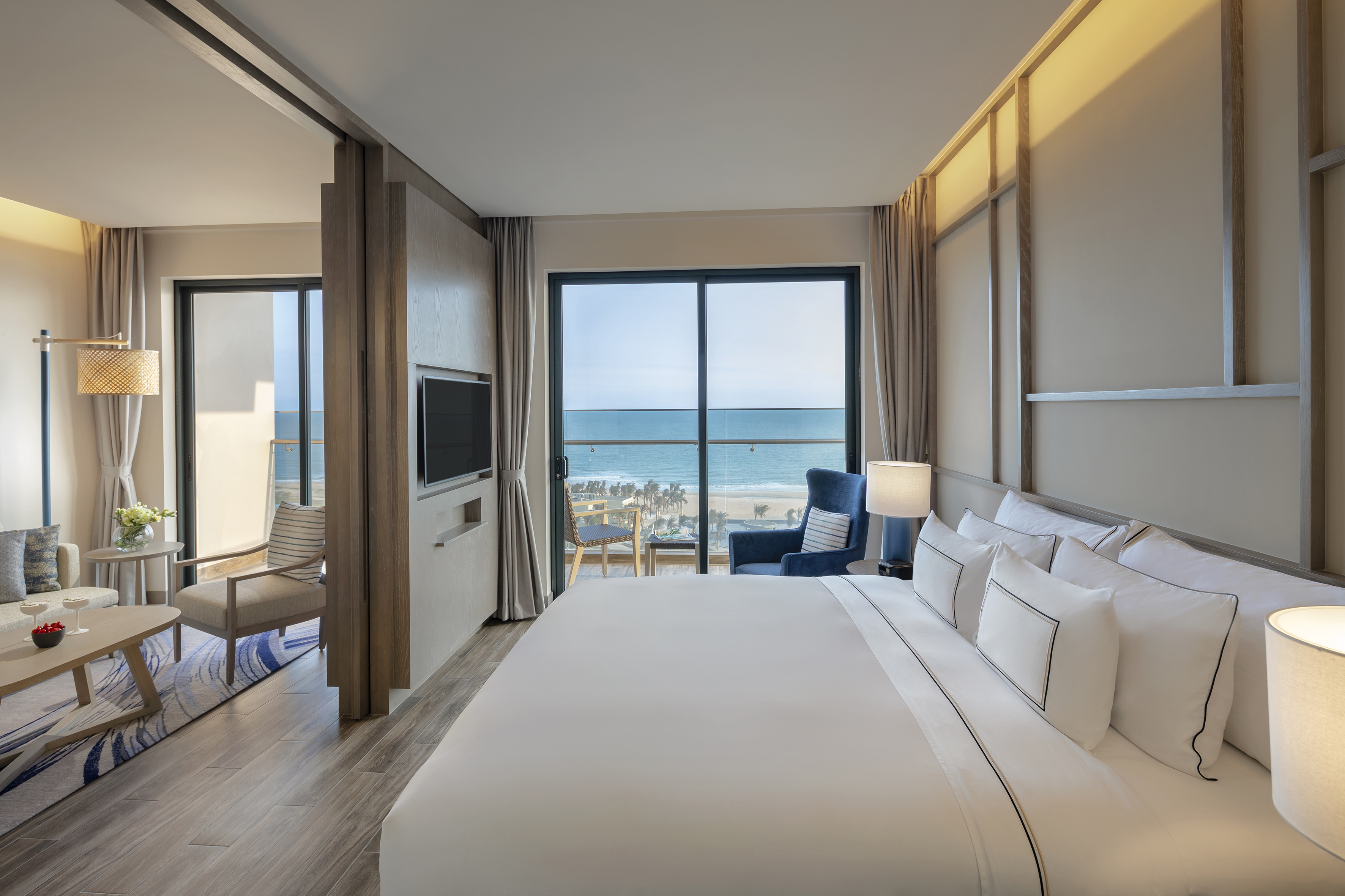 Modern hotel suite with beach and ocean view balcony, bed, and living room.
