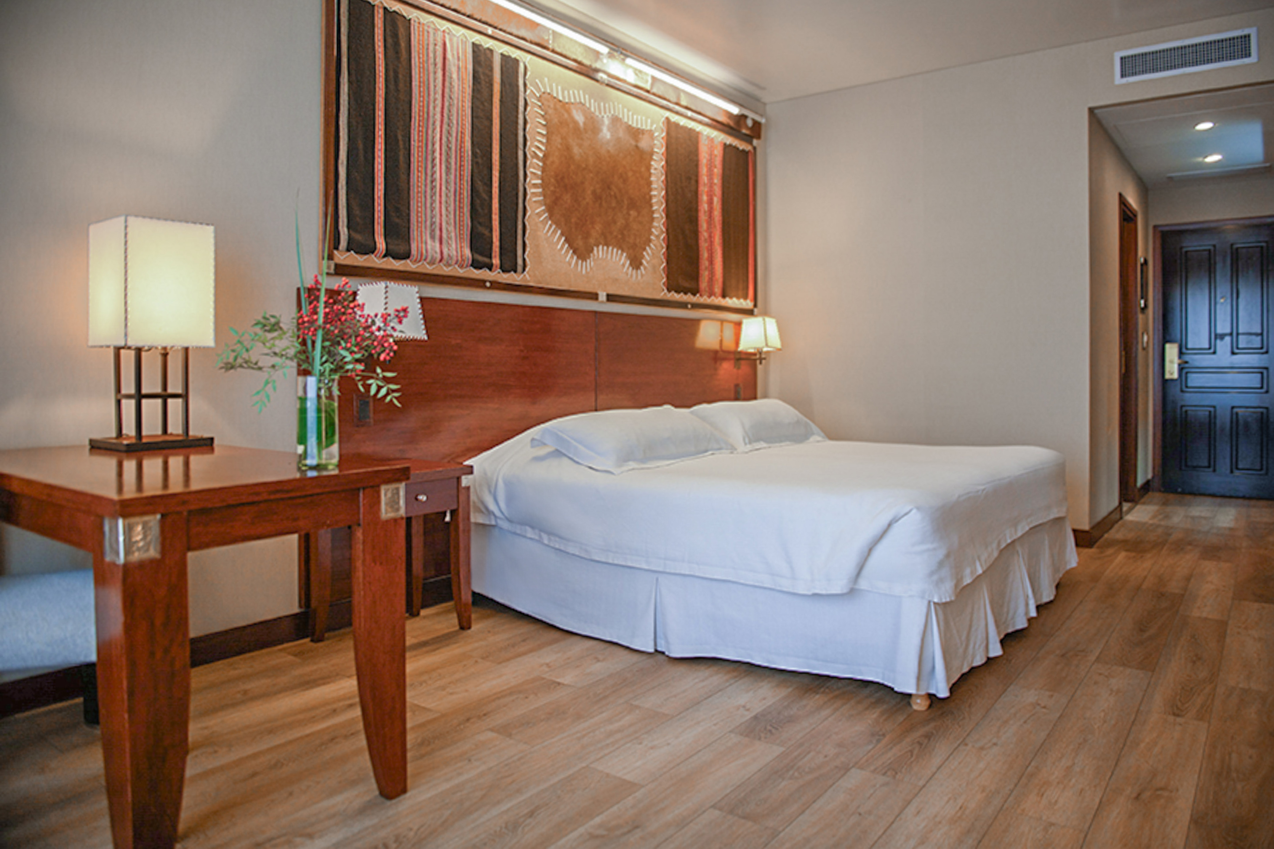 Hotel bedroom with queen bed, wooden furniture, and patterned wall decor.