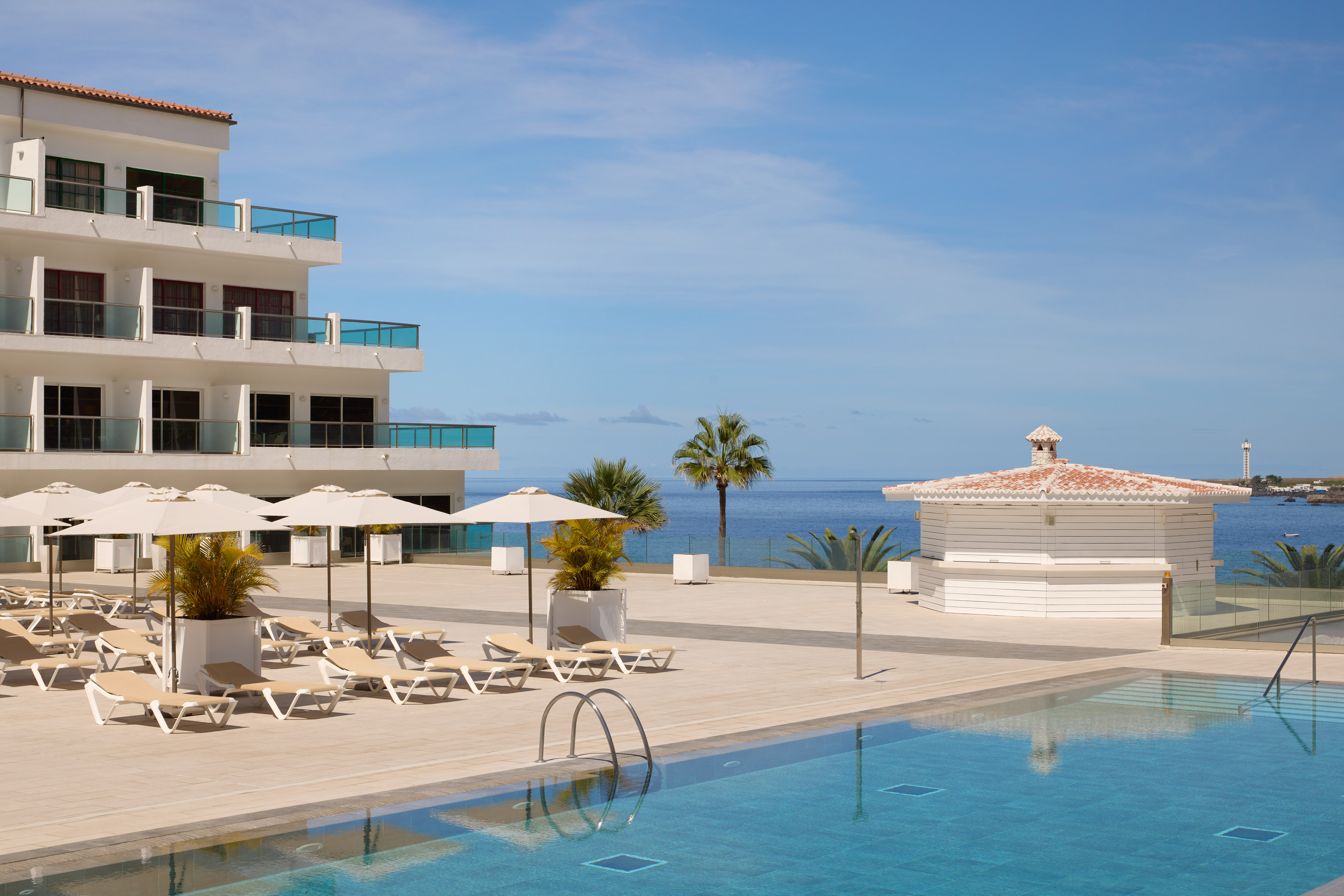 Modern hotel pool with sun loungers, ocean view, and palm trees.