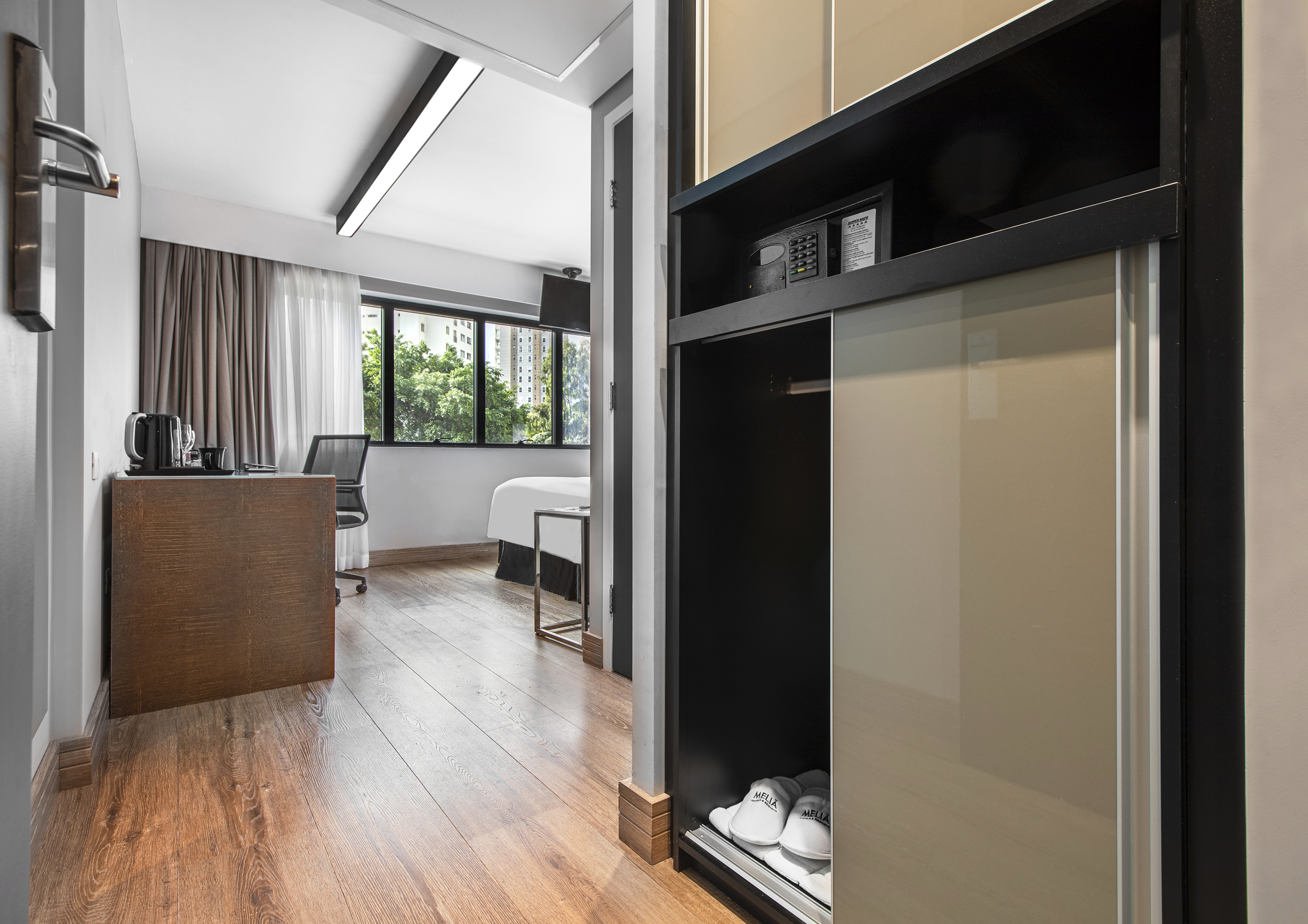 Modern hotel room with wooden floor, desk, window, and wardrobe with safe.