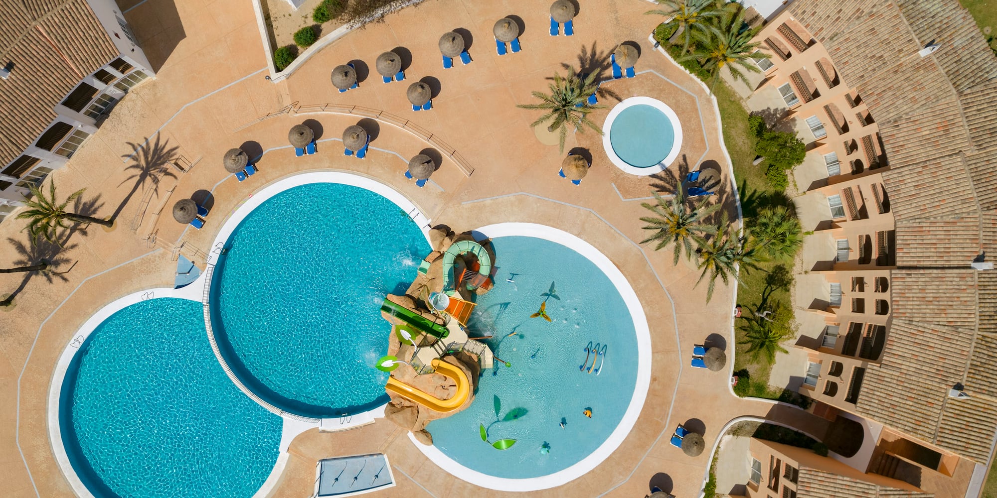 Aerial view: resort pools, kids' water park, sun loungers, palm trees.