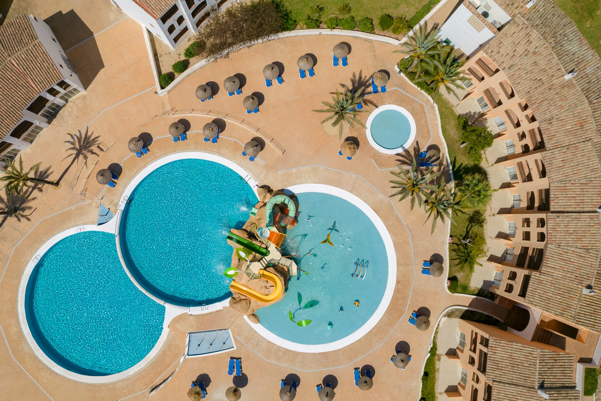 Aerial view of a resort pool area with water park, sunbeds, and palm trees.