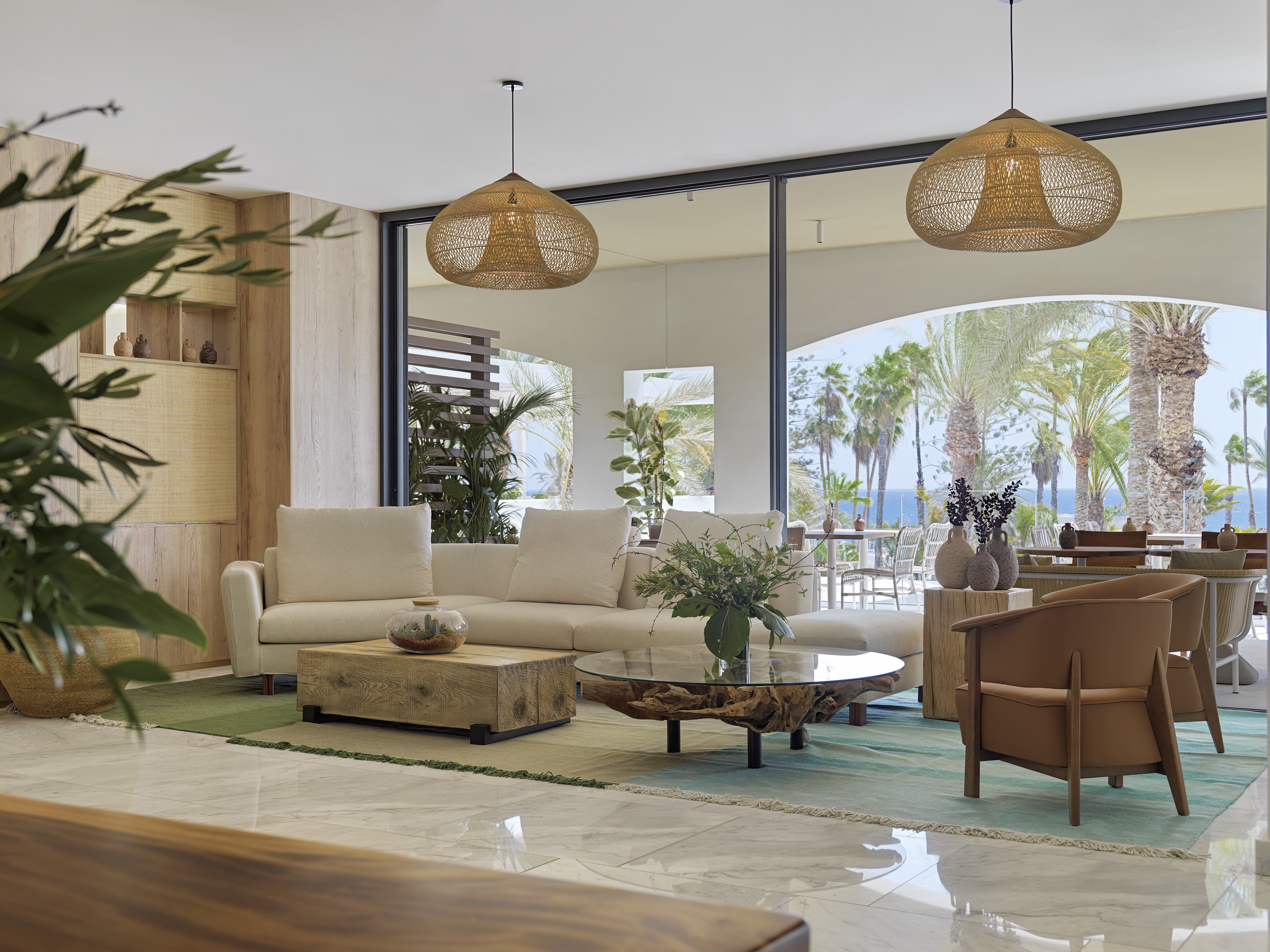 Coastal lounge with ocean view, palm trees, light sofa, and natural wood tables.