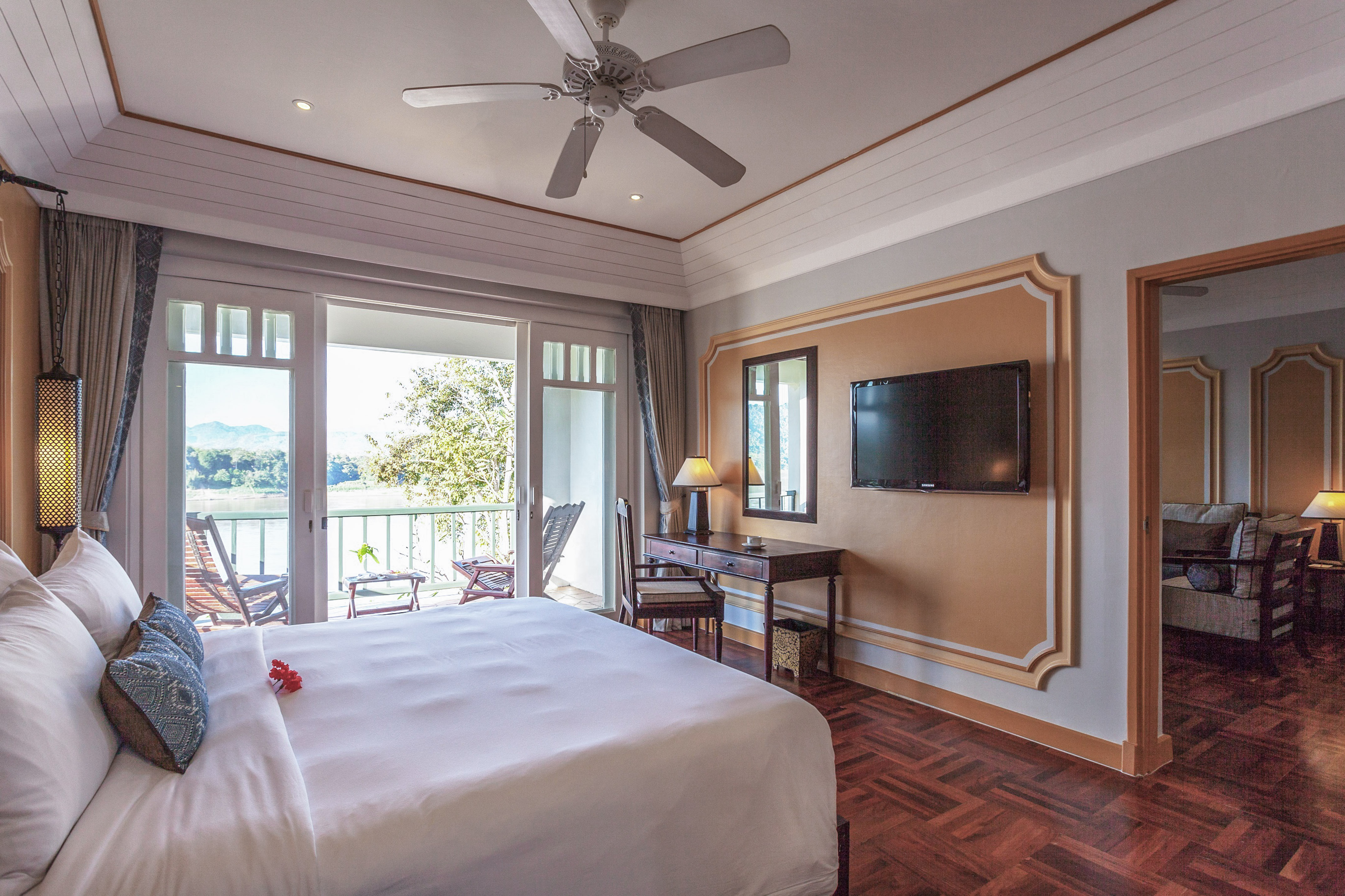 Luxurious hotel bedroom with white bed, river view balcony, and ceiling fan.
