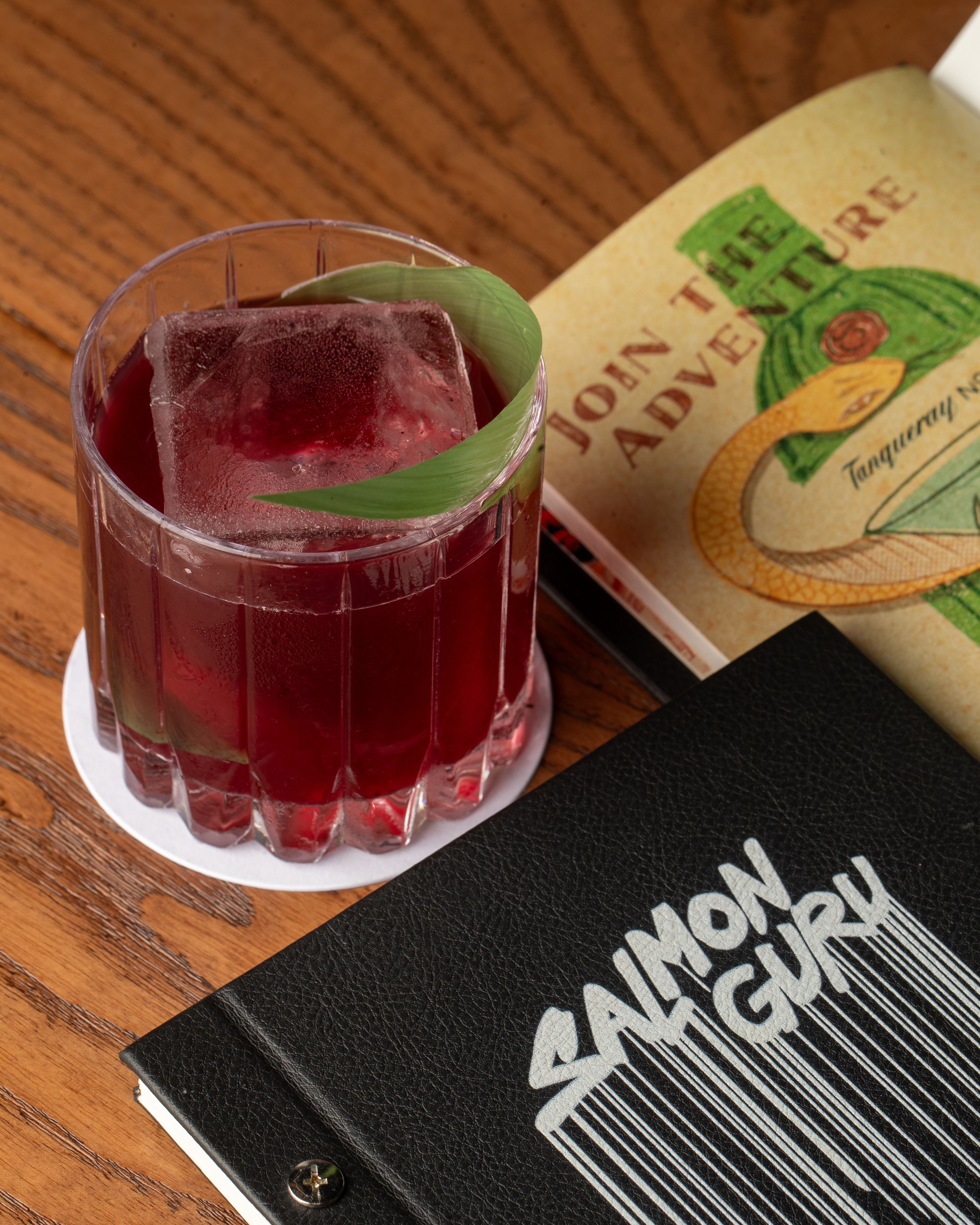 Red cocktail with large ice and leaf garnish, plus Saimon Guru menu on wood.