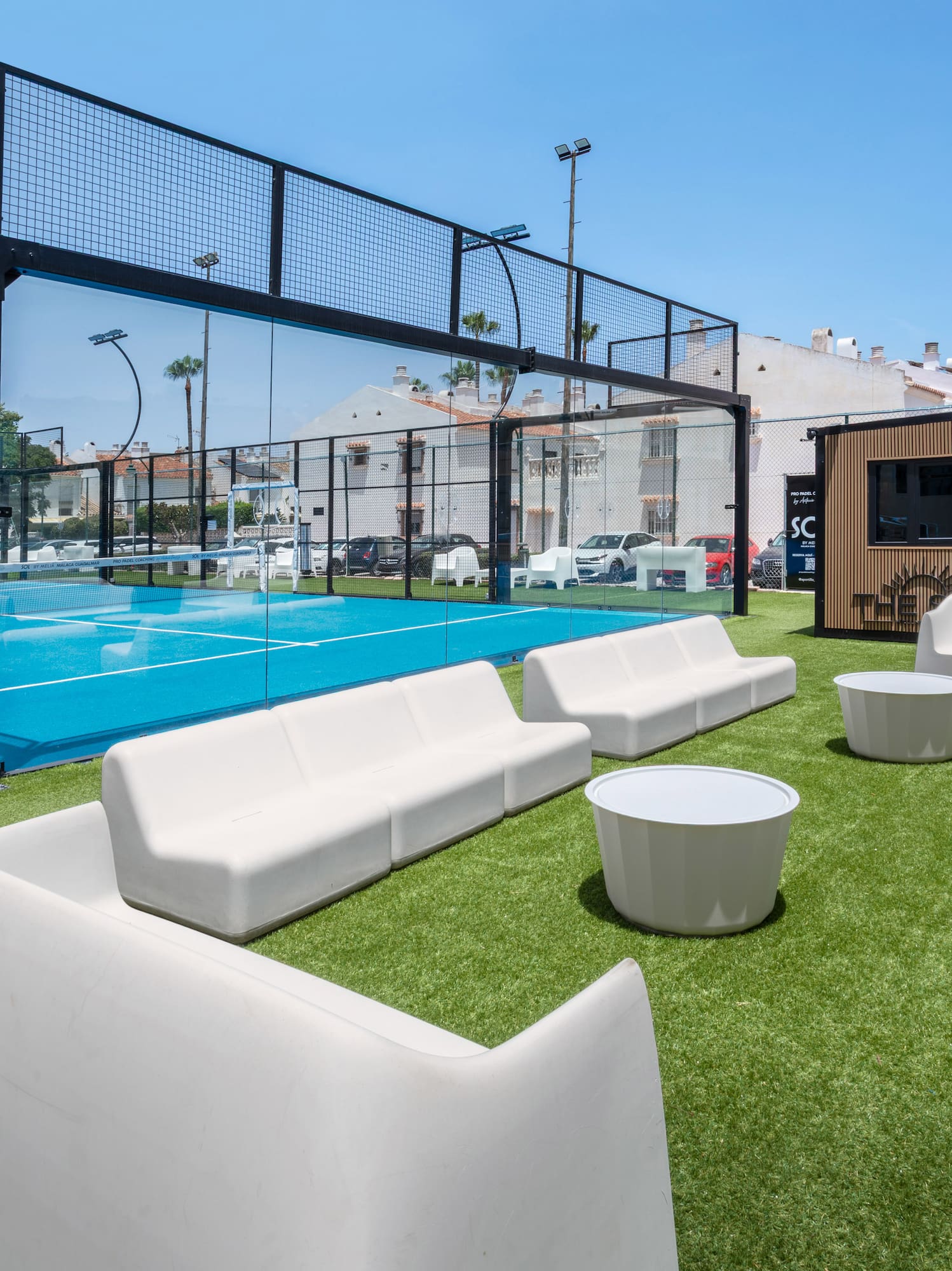 Blue padel court with artificial turf lounge area, white outdoor sofas.
