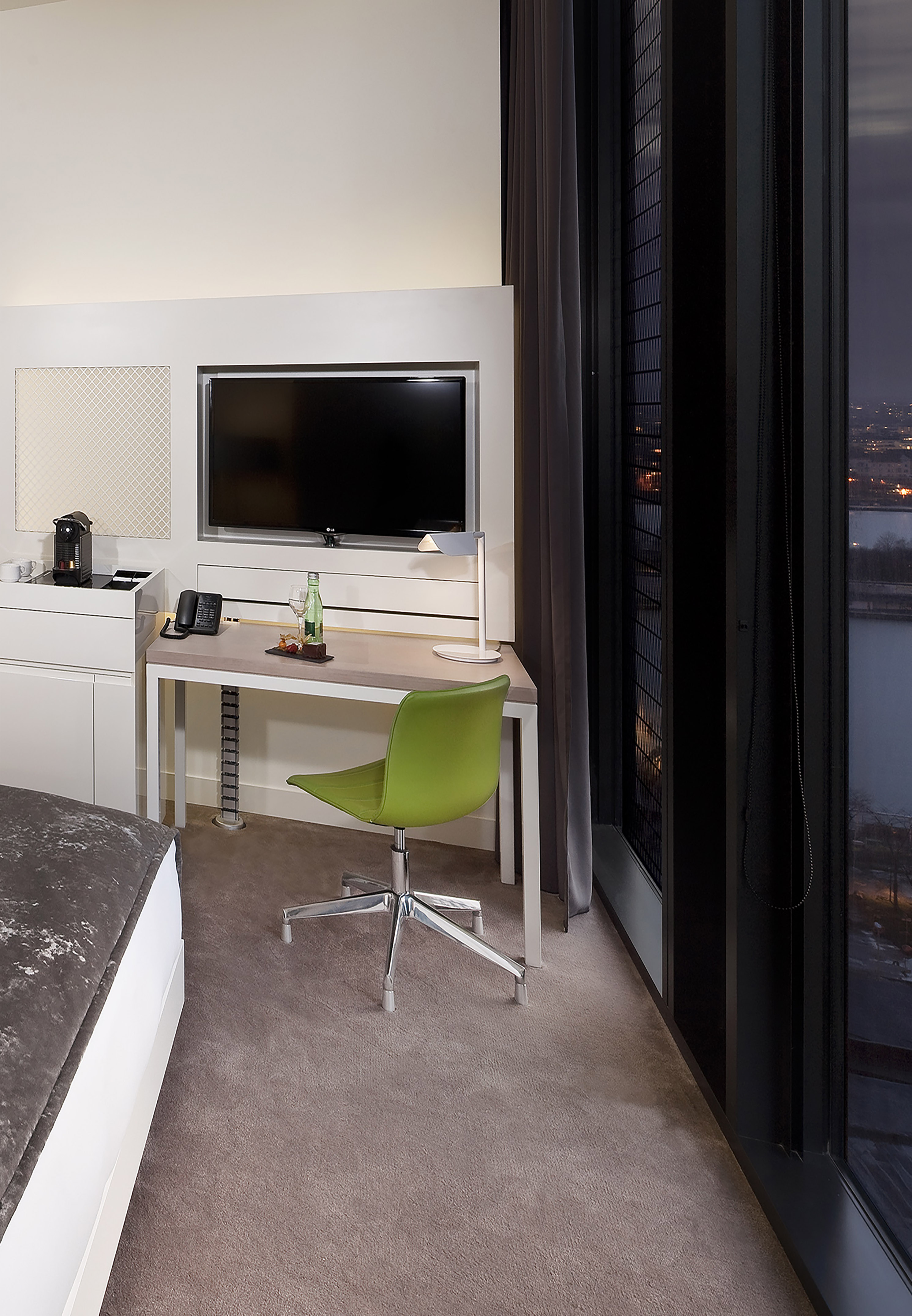 Modern hotel room with work desk, green chair, TV, and city night view.
