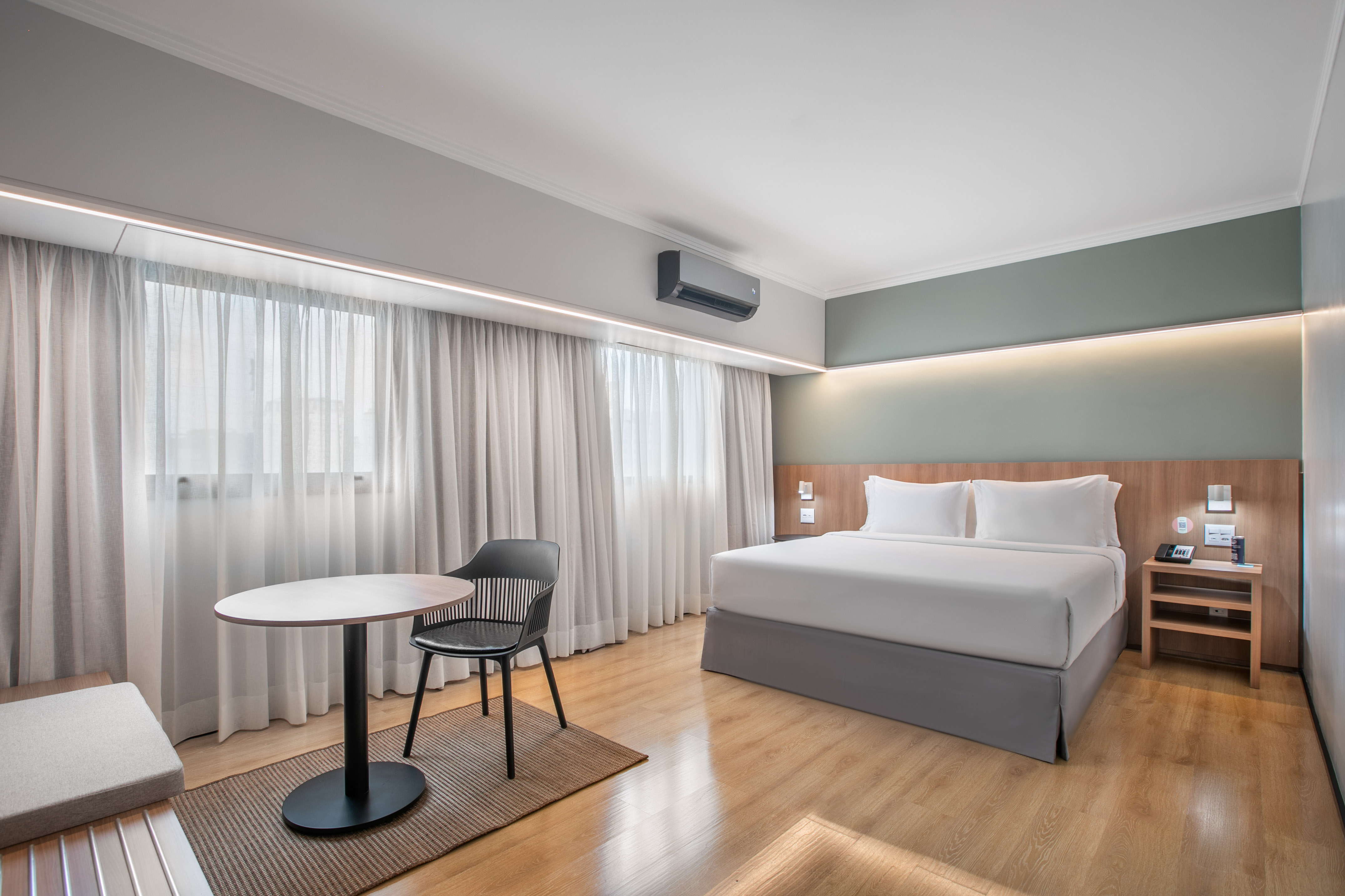 Modern hotel bedroom with king bed, wooden floor, AC, and sheer curtains.
