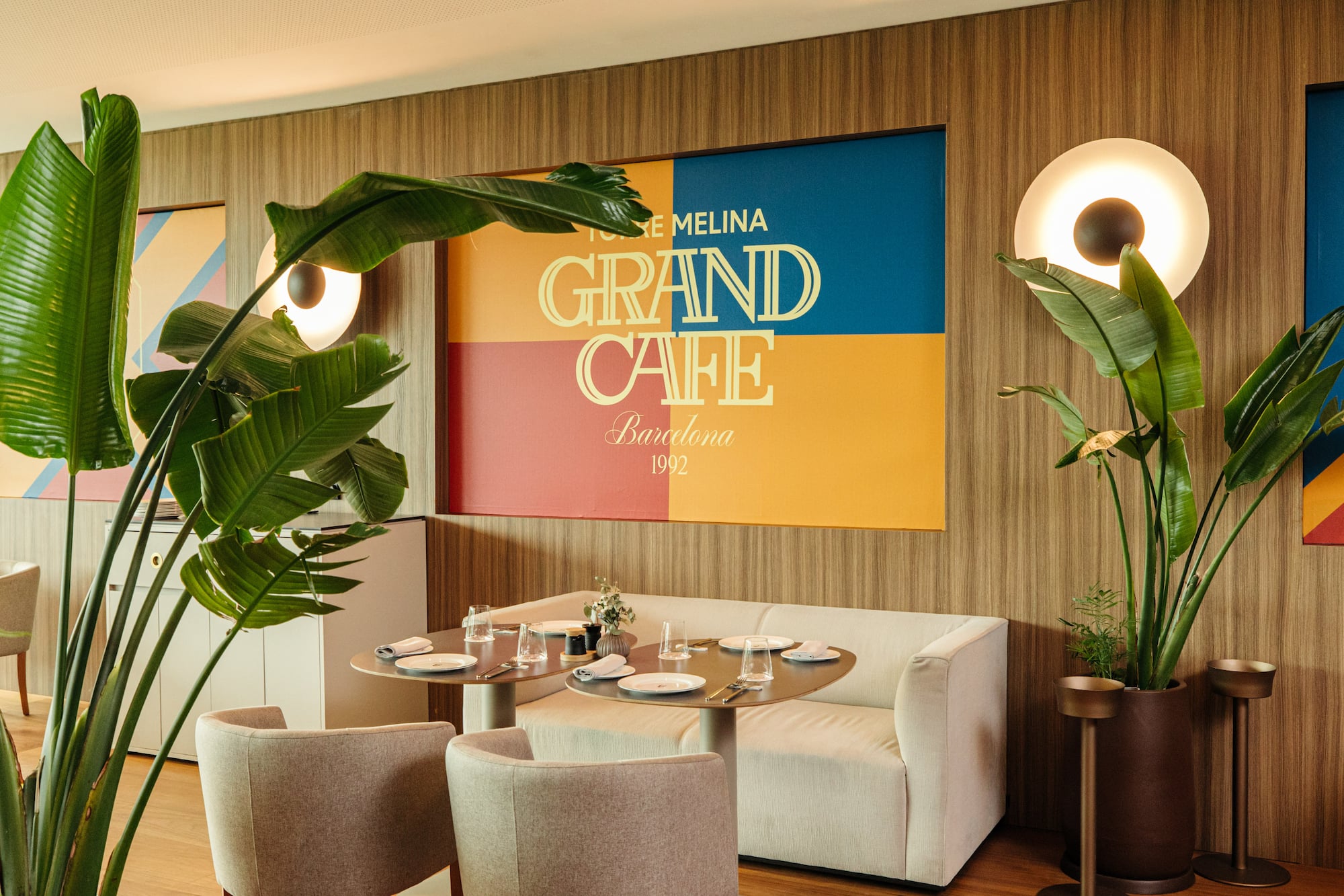 Modern Grand Cafe Barcelona interior with colorful sign, plants, and dining tables.